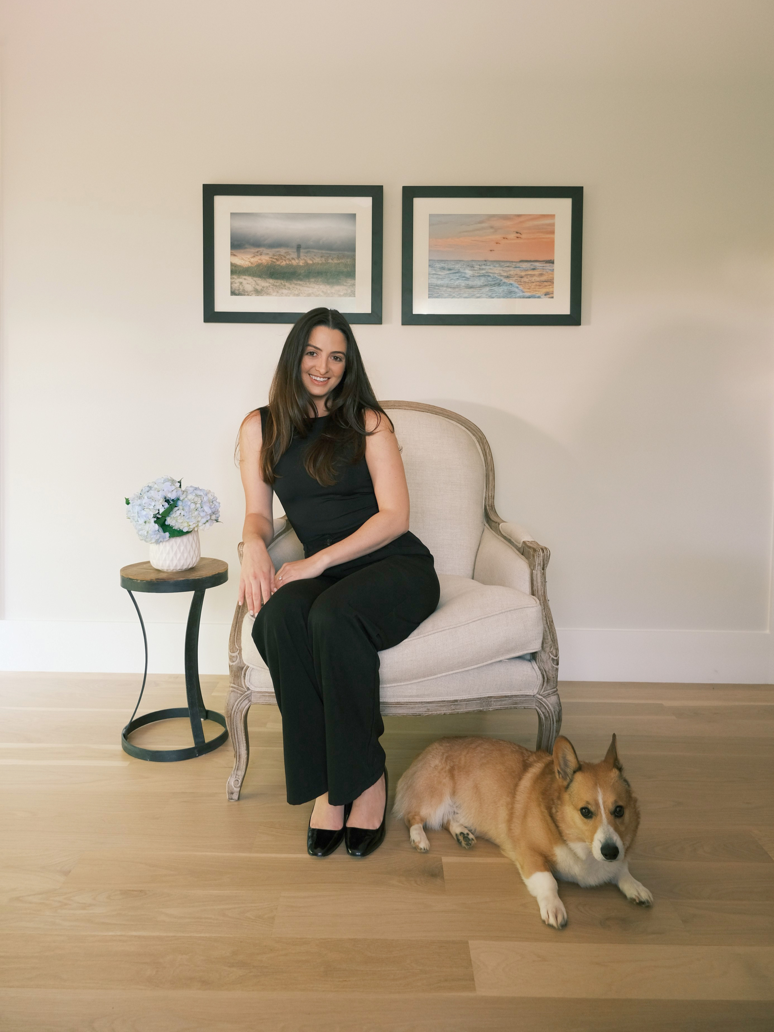 A woman in a black outfit sitting on a vintage-style armchair with a small dog lying on the floor beside her. There is a side table with a white ceramic vase of purple and white flowers. Two framed landscape photographs are hung above her on a cream-colored wall.