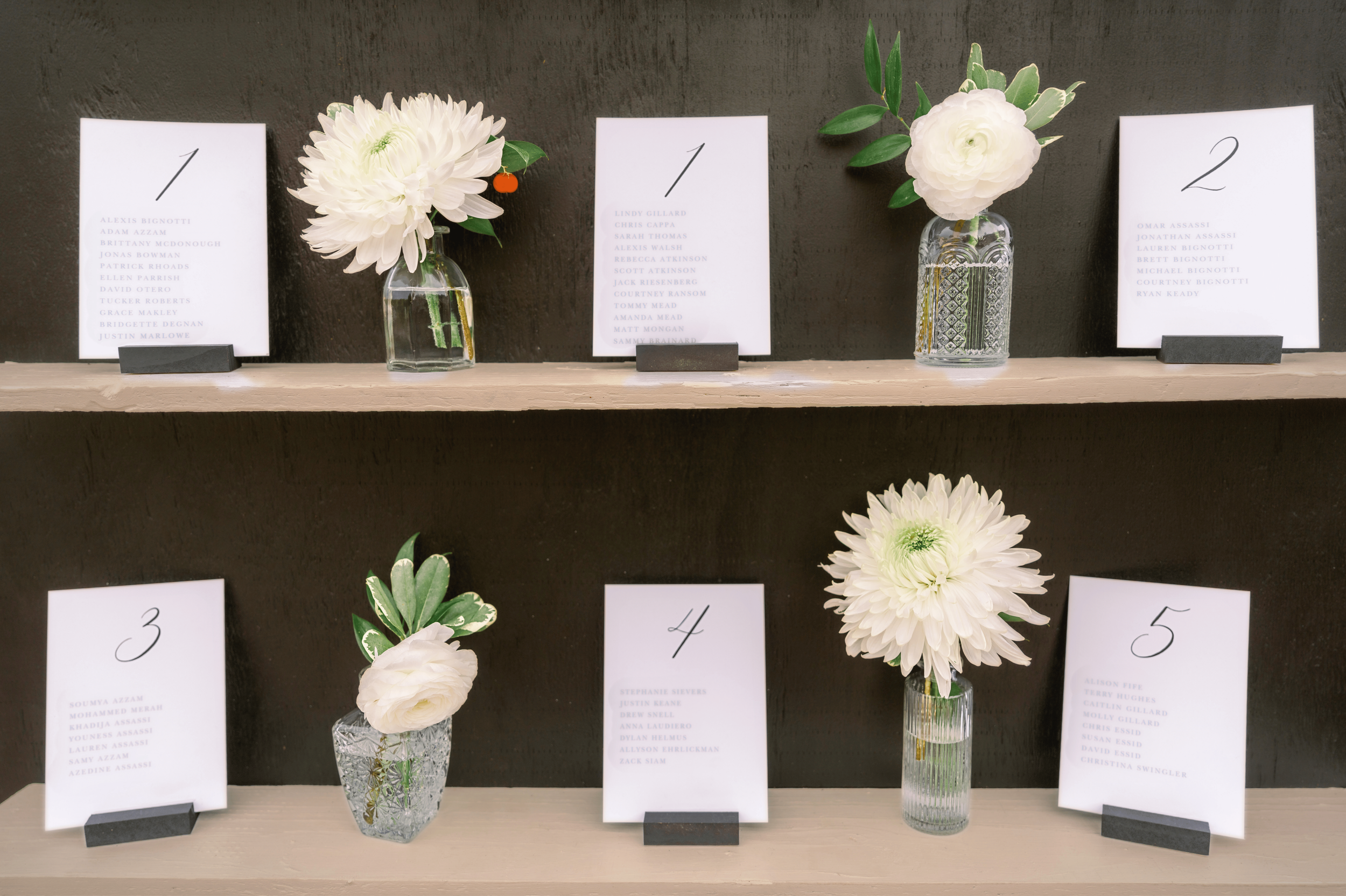Three small vases with white flowers and greenery, each placed next to a sheet of paper with a list of names, on a wooden surface against a black wall.