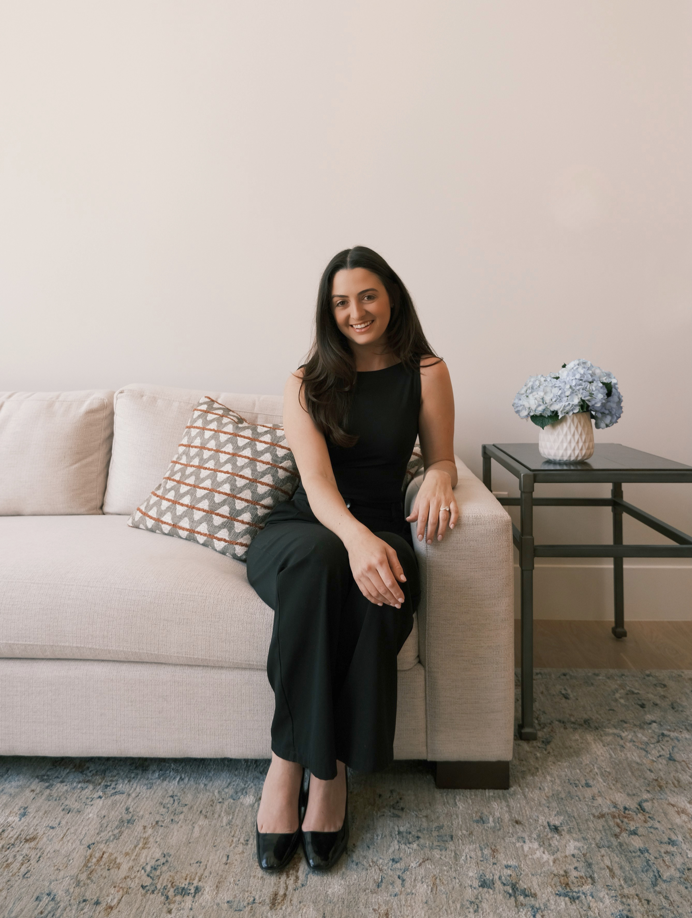 A woman with long dark hair sitting on a beige sofa, smiling, wearing a sleeveless black dress, with her legs crossed at the ankles, next to a side table with a white vase of blue hydrangeas, in a room with a plain wall and a patterned cushion.