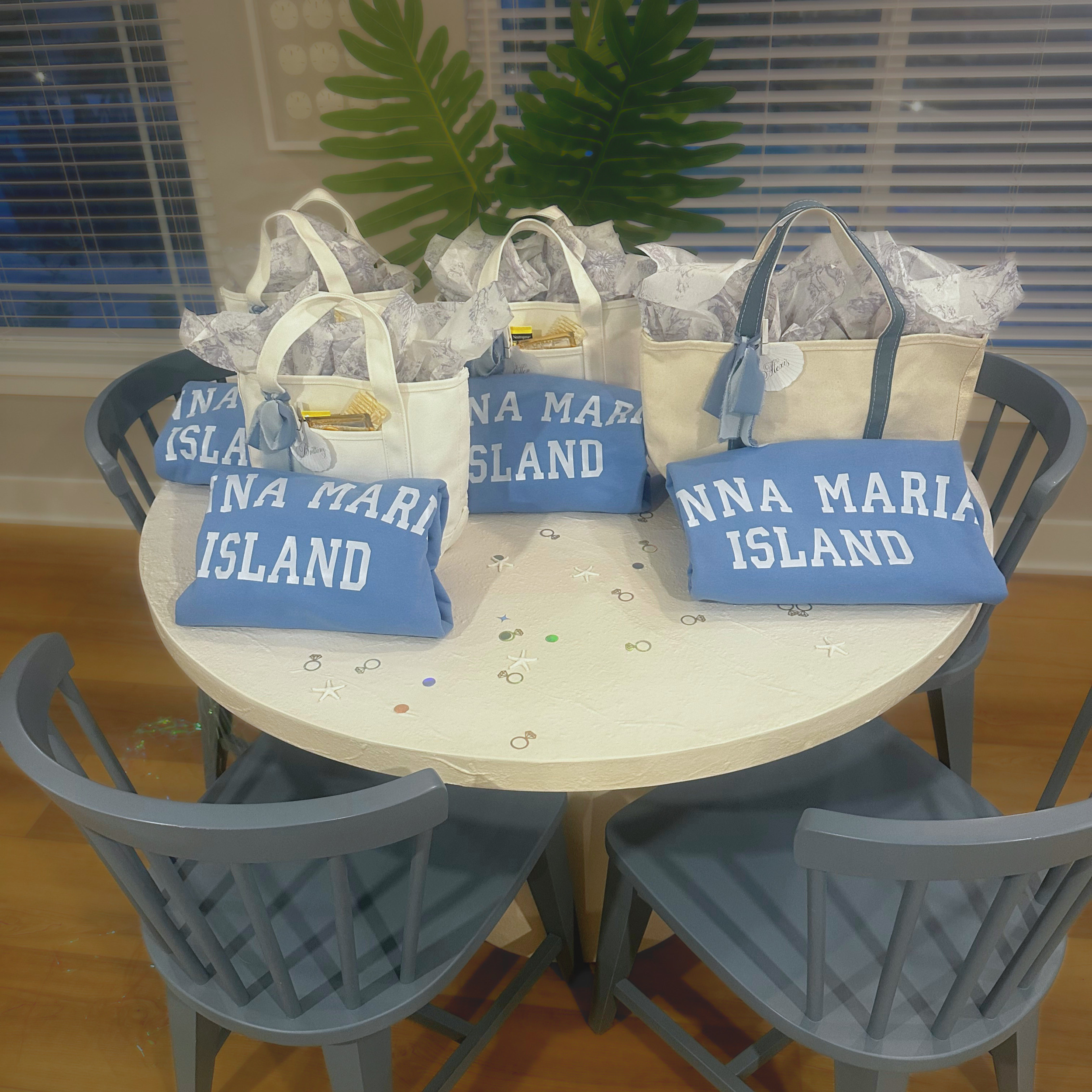 Multiple light blue tote bags with white handles and the words 'NNA MARIA ISLAND' written on them, placed on a white table with starfish confetti, under a large leafy green plant, with window blinds in the background.