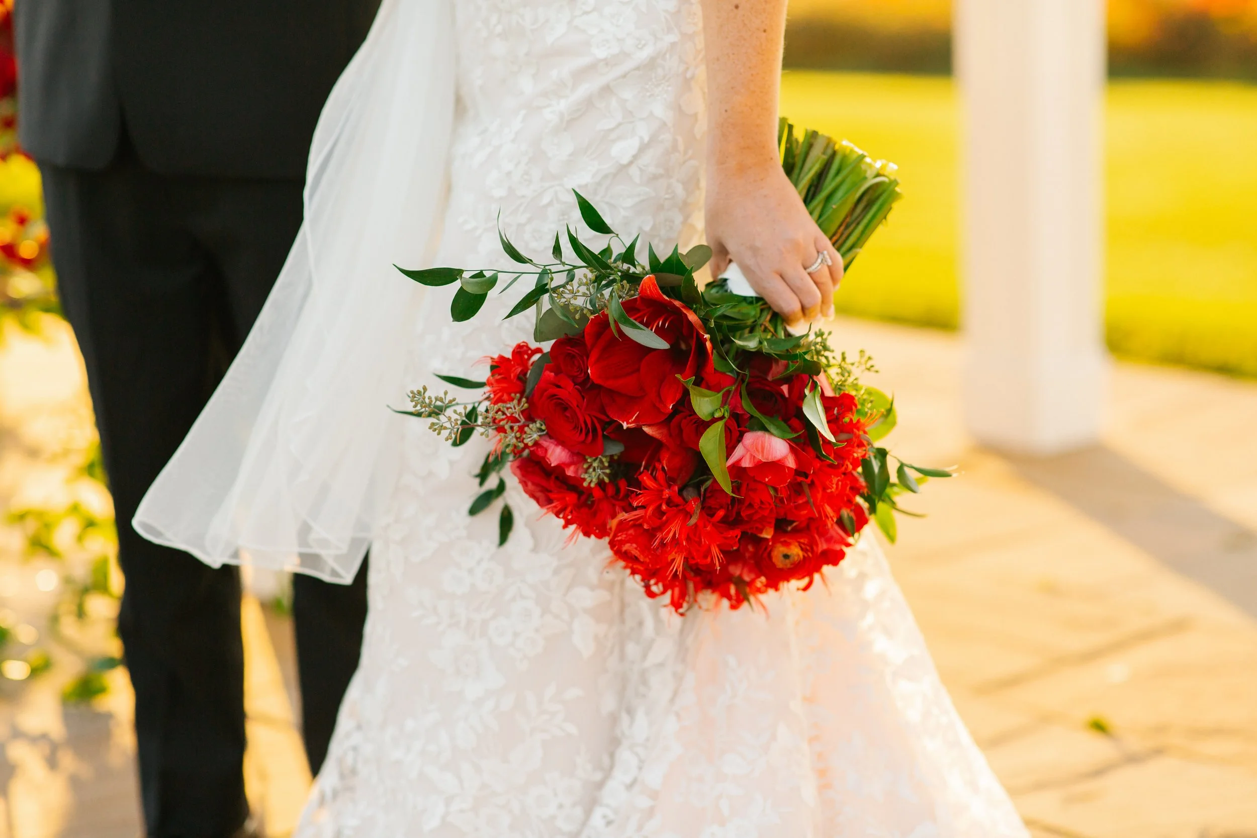 A bride in a white wedding dress holding a bouquet of red flowers, standing outdoors with sunlight, with a groom in a black suit partially visible beside her.