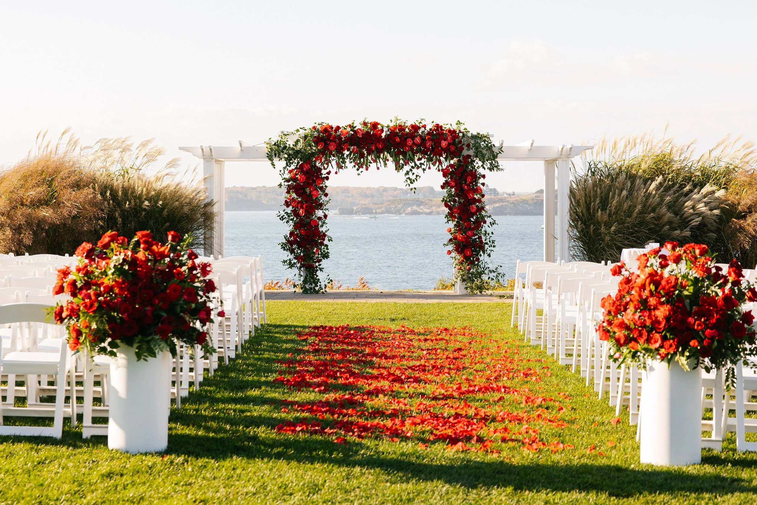 Outdoor wedding ceremony setup with white chairs, floral arch, red flower arrangements, and rose petal aisle by water with island in the background.