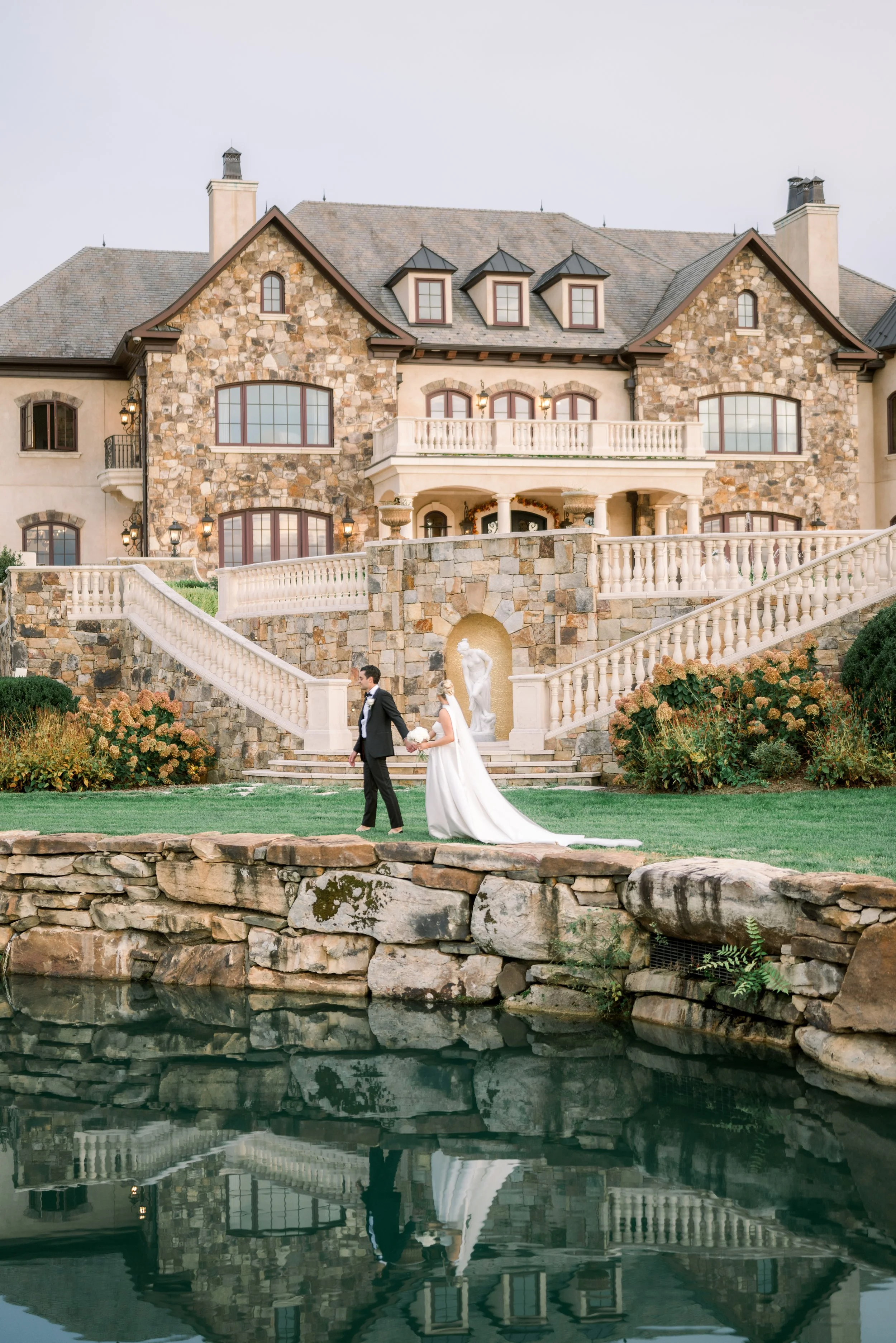 A bride and groom holding hands and walking along a grassy area in front of a large stone mansion during their wedding.