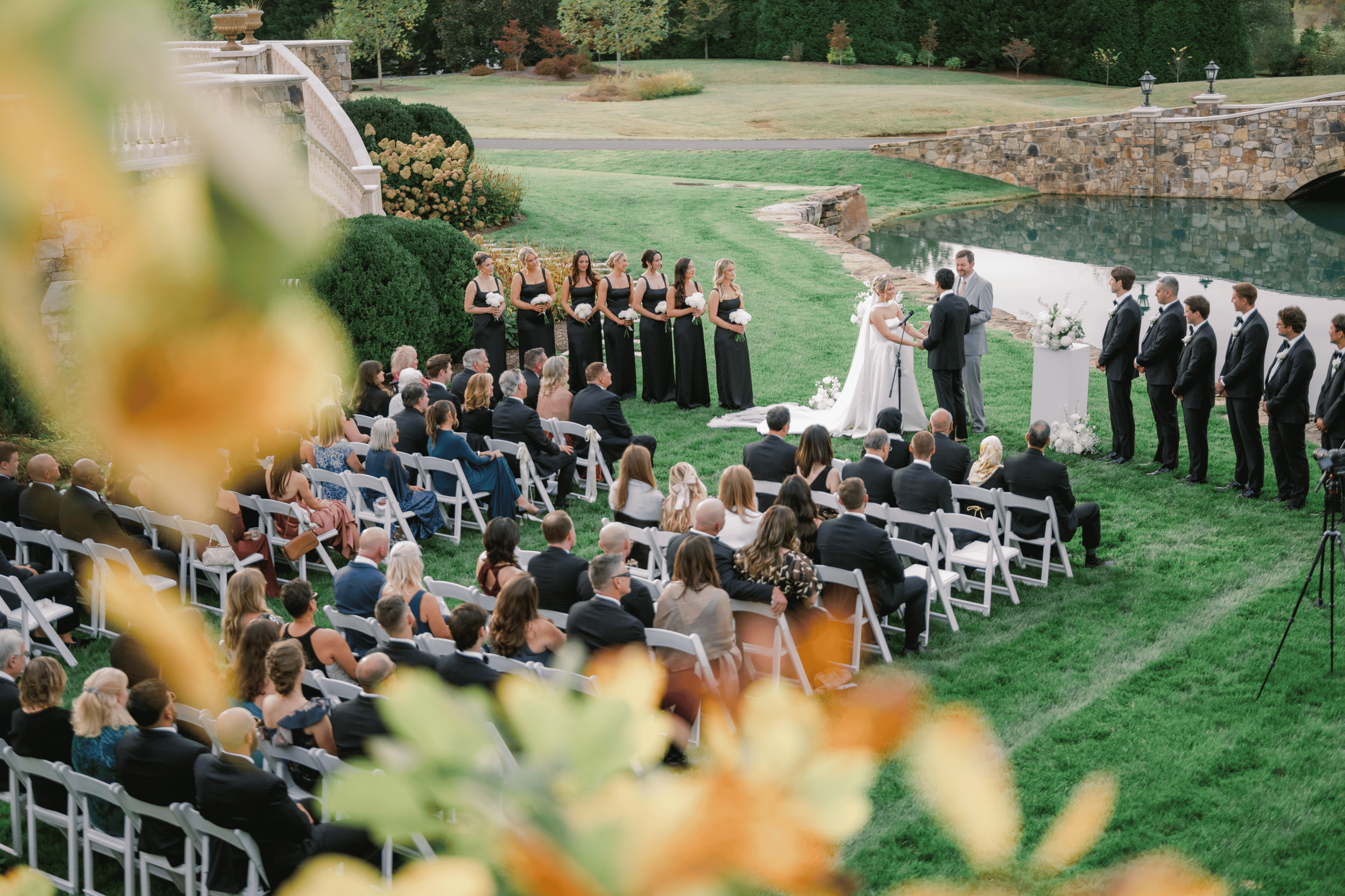 A wedding ceremony taking place outdoors on a grassy lawn by a pond, with guests seated in rows and a bride and groom exchanging vows in front of an officiant, surrounded by bridesmaids and groomsmen, with scenic trees and a stone bridge in the background.