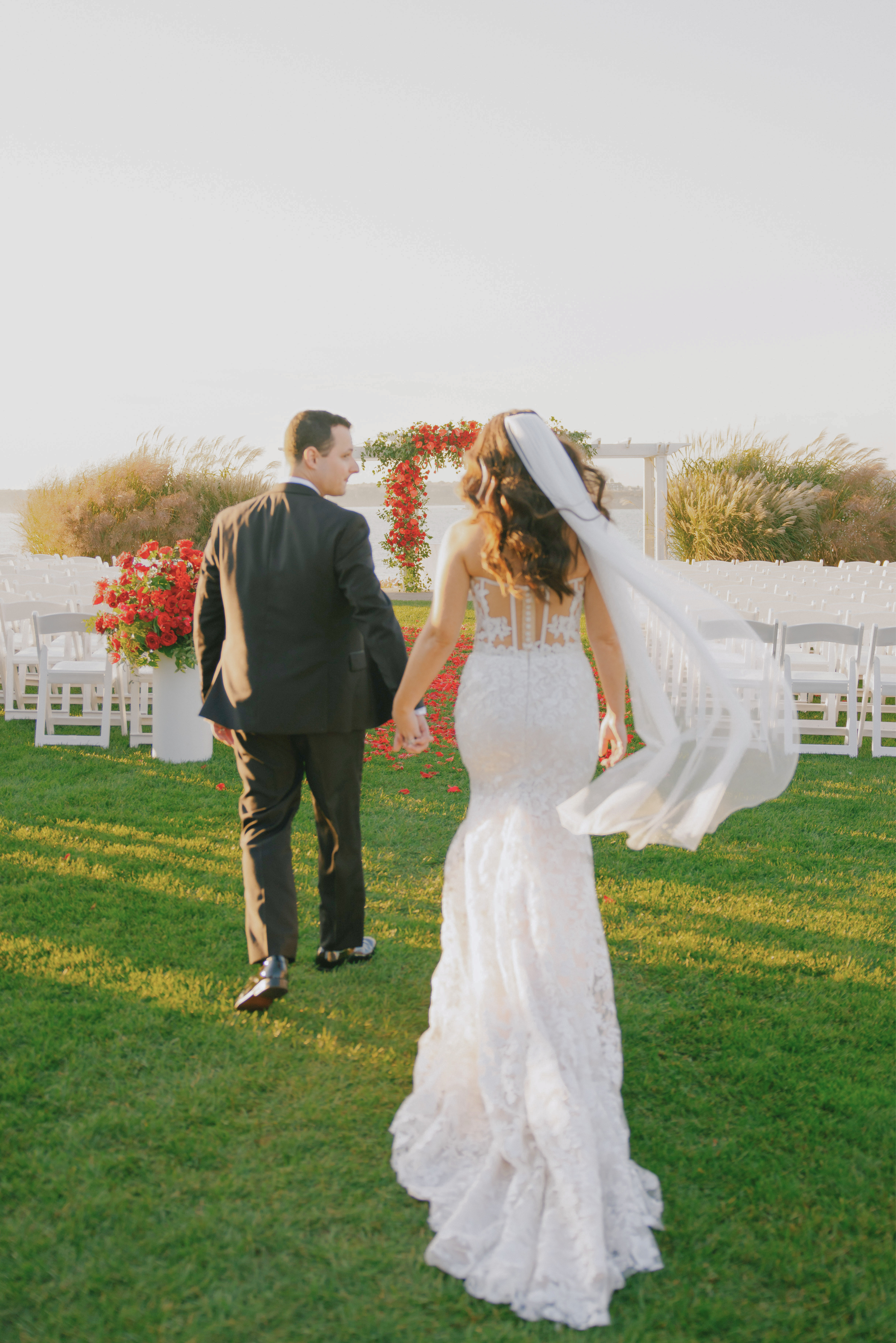 A bride and groom holding hands and walking down the aisle at an outdoor wedding ceremony during sunset, with chairs and floral decorations in the background.