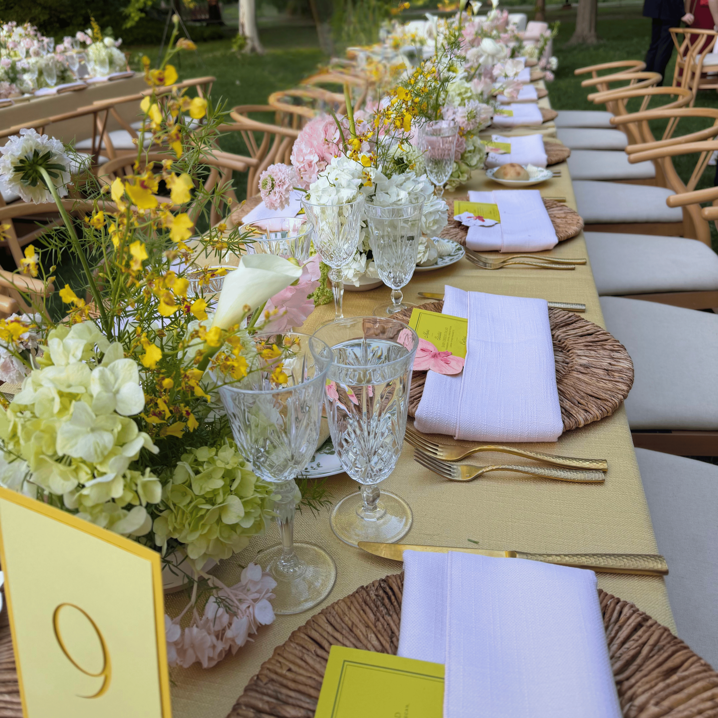A long outdoor dining table decorated with floral arrangements, white napkins, and glassware, set for a celebration in a garden area.