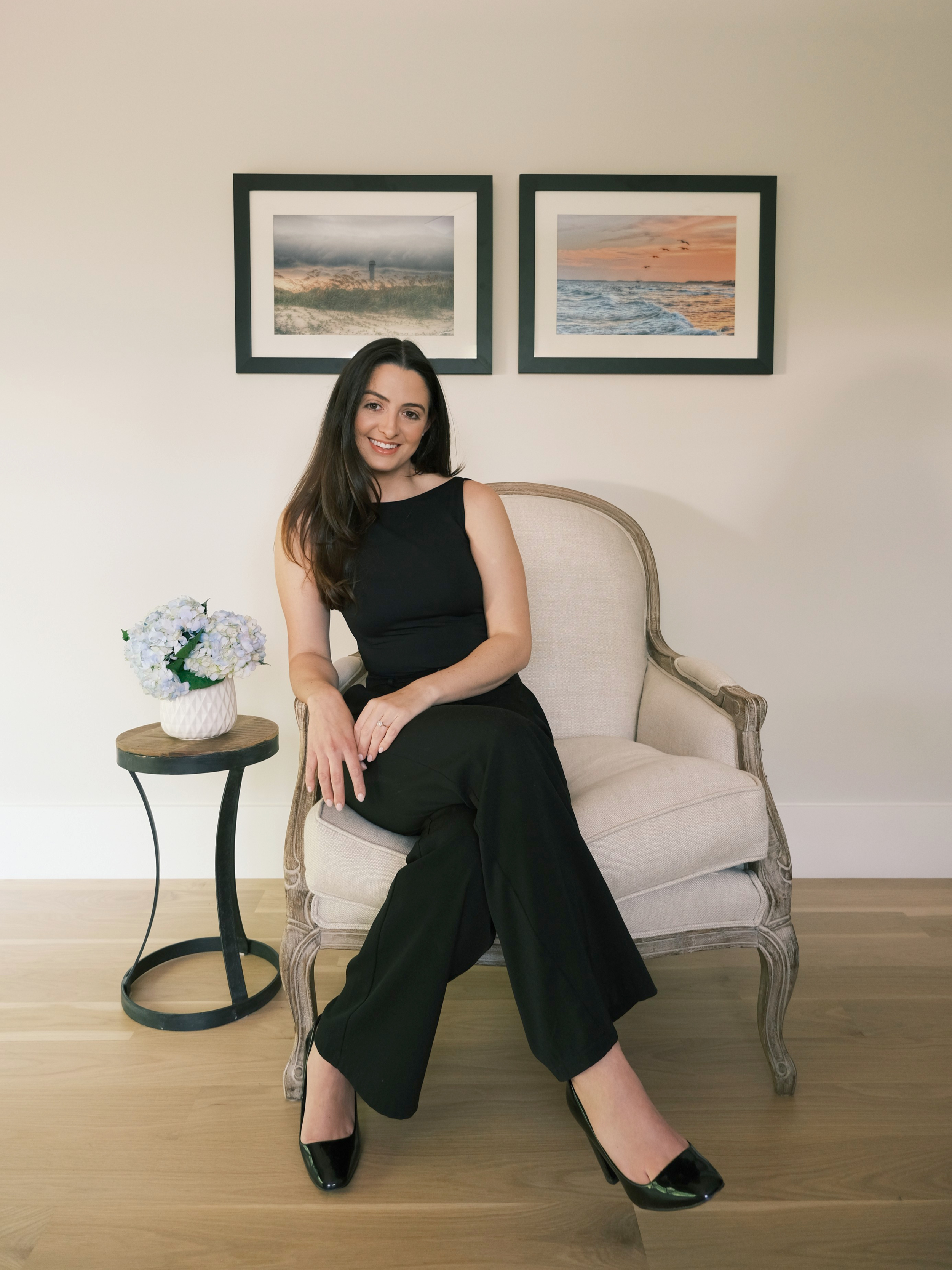 A woman with long dark hair, dressed in a black sleeveless top, black pants, and black heels, is sitting on a beige vintage-style armchair. She is smiling and has her hands resting on her lap. Next to the chair is a small round side table with a white vase of light purple flowers. Behind her, a wall features two framed landscape photographs, one of a cloudy sky over a shoreline and the other of a sunset over the ocean. The floor is light wood, and the wall is plain white.