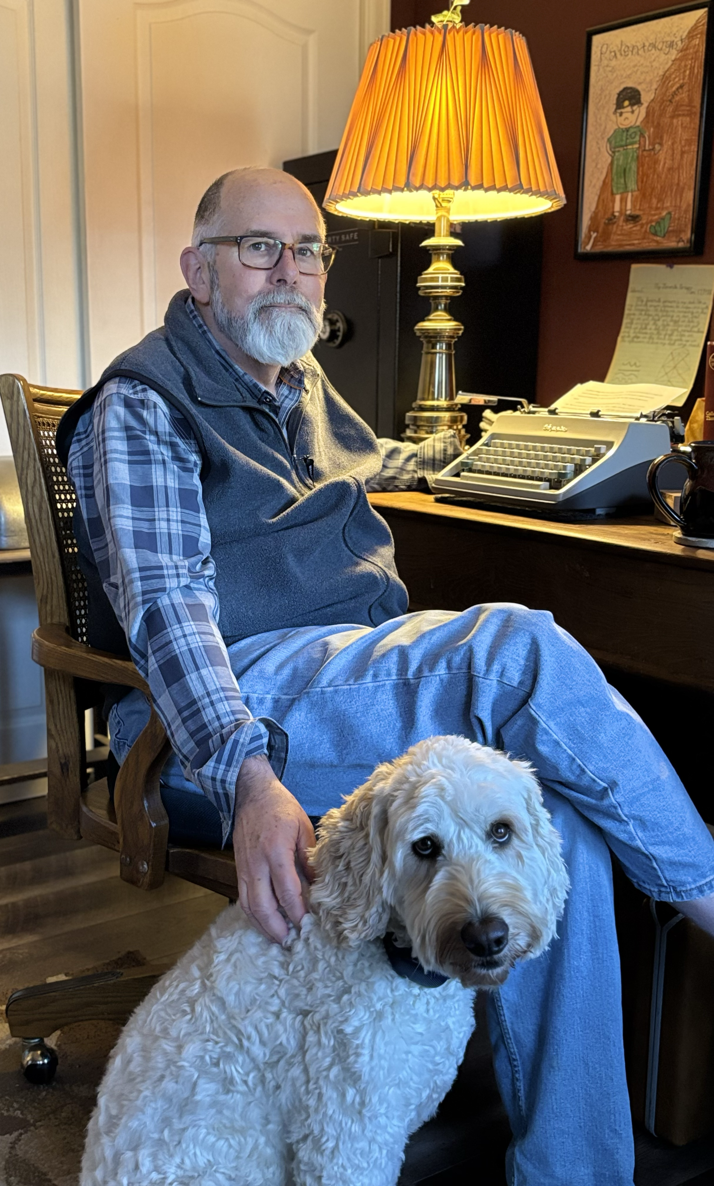 An older man with glasses and a beard sitting in a wooden chair, petting a large, fluffy, light-colored dog in a cozy room with a desk, a lit table lamp, a typewriter, a safe, and artwork on the wall.