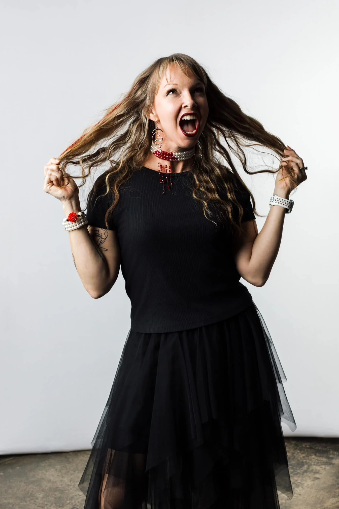 A woman with long, wavy blonde hair wearing a black dress, red lipstick, and pearl jewelry, standing against a plain white background, holding her hair with both hands, and showing an excited or shouting expression.