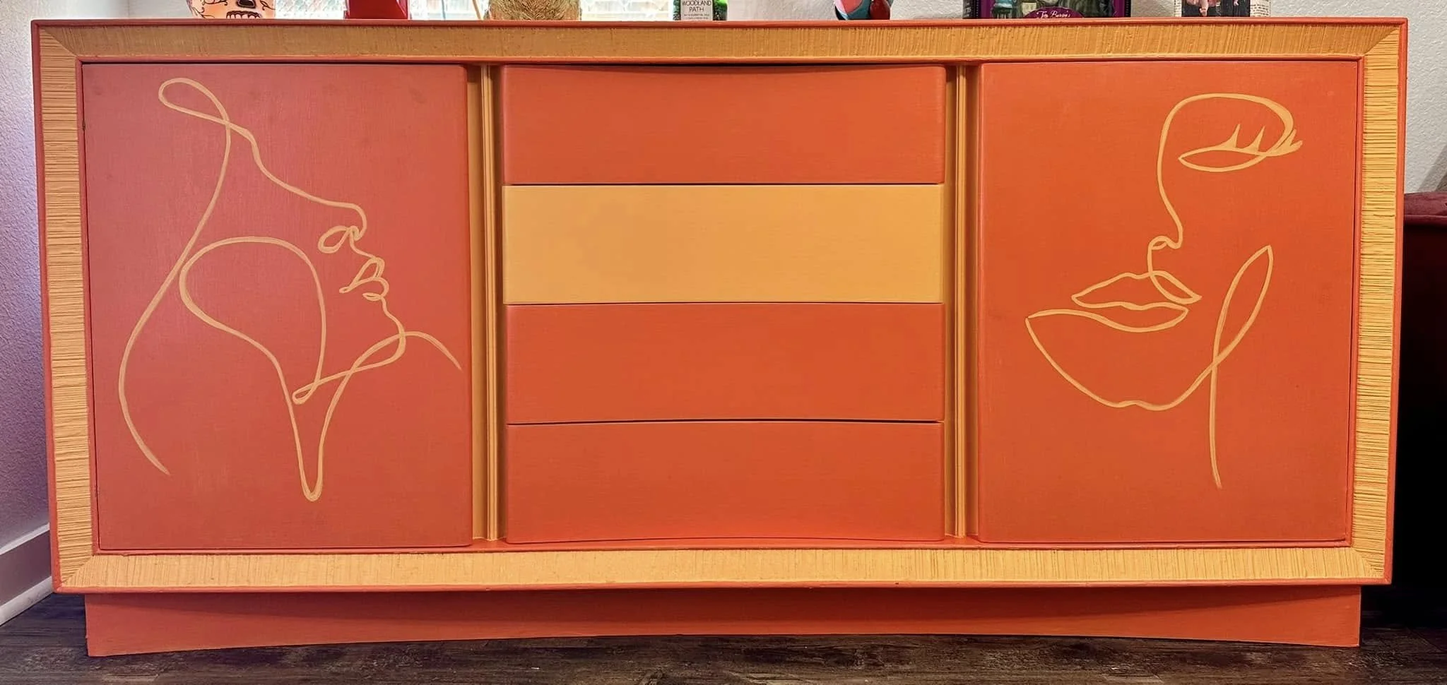 A wooden cabinet with orange and yellow painted doors, featuring minimalist line art of two faces touching. One face is on the left with hair, and the other on the right with a more pronounced nose and lips.