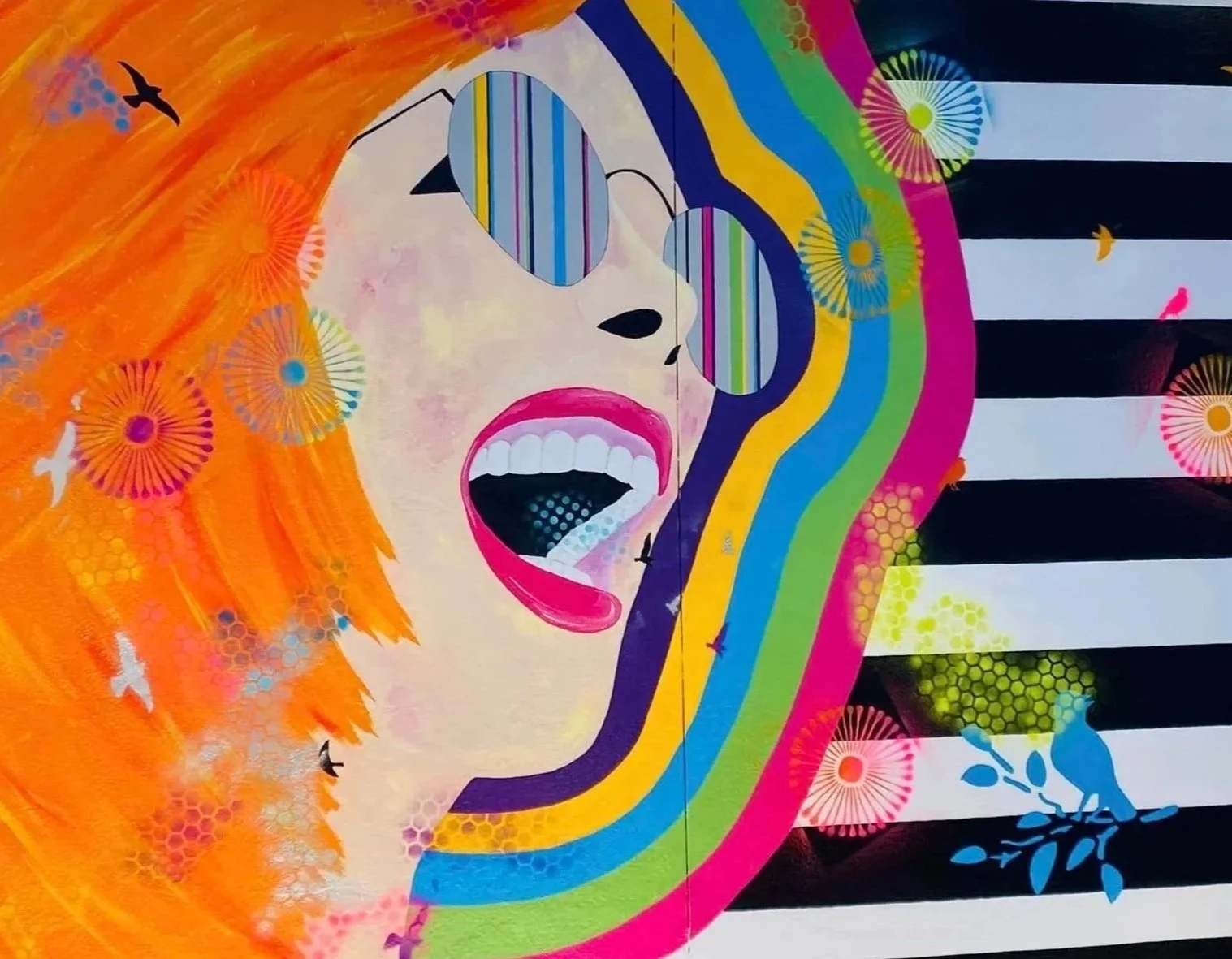Colorful mural of a woman with orange hair, wearing glasses, singing with her mouth open, surrounded by vibrant abstract patterns, flowers, birds, and geometric shapes.