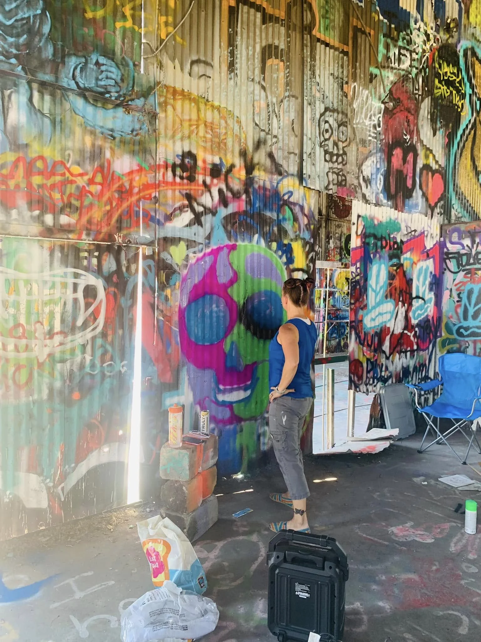 A person with a bun hairstyle, wearing a blue sleeveless top, standing in front of a colorful graffiti-covered wall, in an outdoor setting. Art supplies and chairs are visible around.