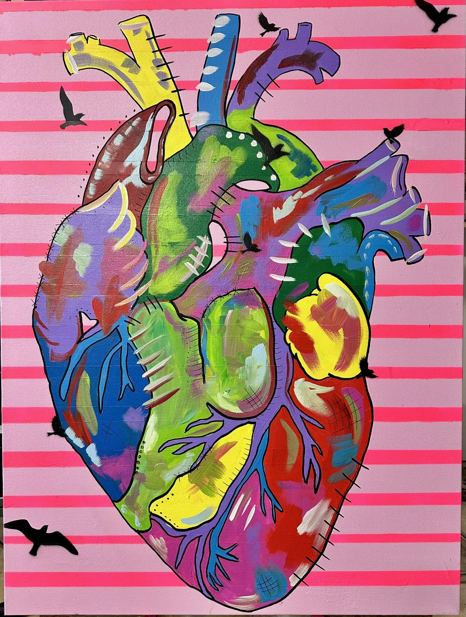 Colorful street art painting of a human heart with various sections painted in bright colors like yellow, green, purple, blue, red, and pink, set against a background of pink and red horizontal stripes, with black birds flying around.