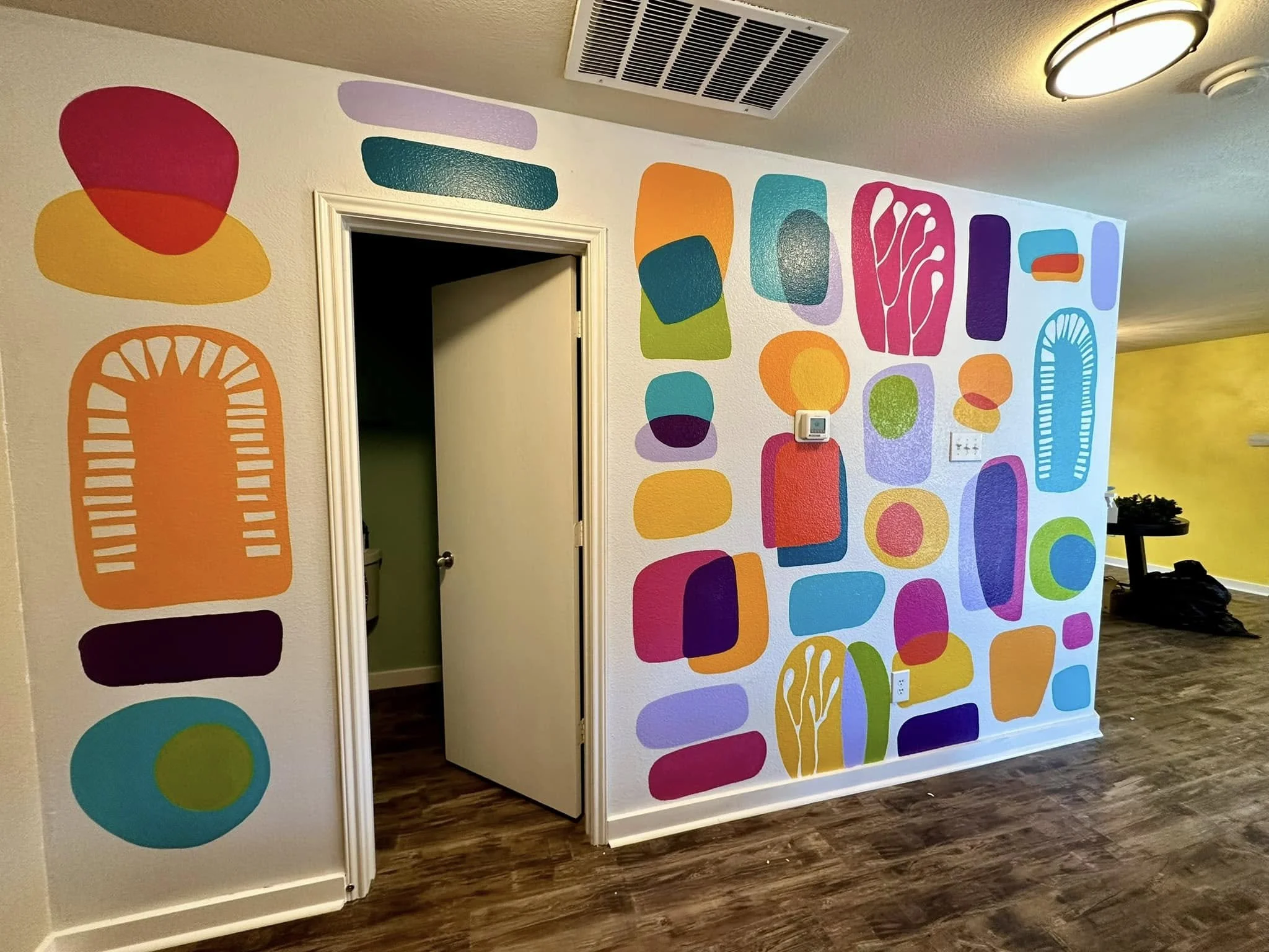 Colorful abstract mural with various shapes and patterns on a white wall in an indoor space.