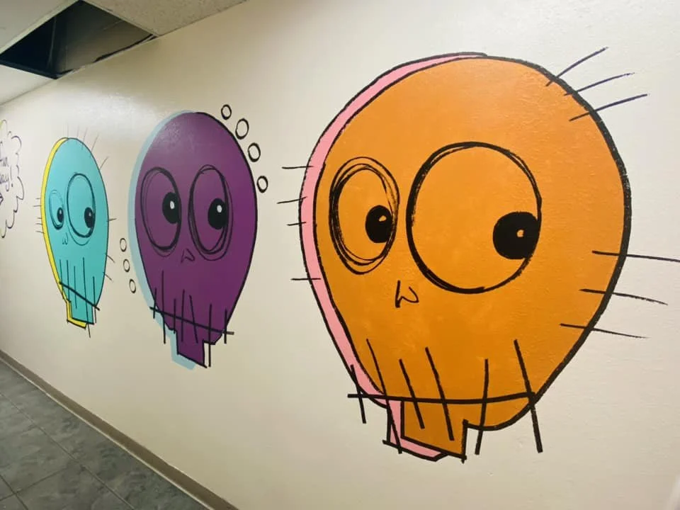 Colorful graffiti of three cartoon-style skulls in yellow, purple, and orange on a white wall.