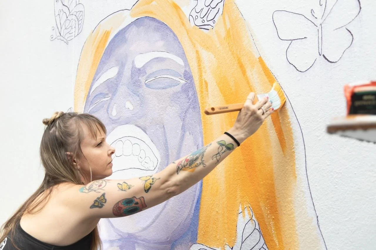 A tattooed woman painting a mural of a crying face with tears and orange hair on a white wall, with outlines of butterflies around.