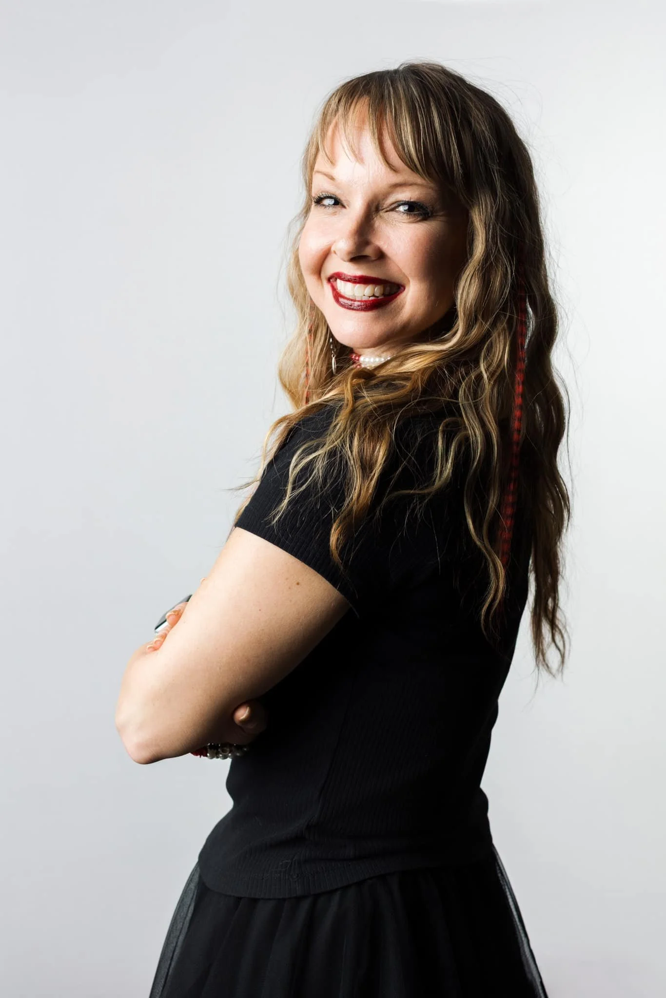 A woman with wavy light brown hair, smiling with teeth visible, wearing a black short-sleeve top, standing with crossed arms against a plain gray background.