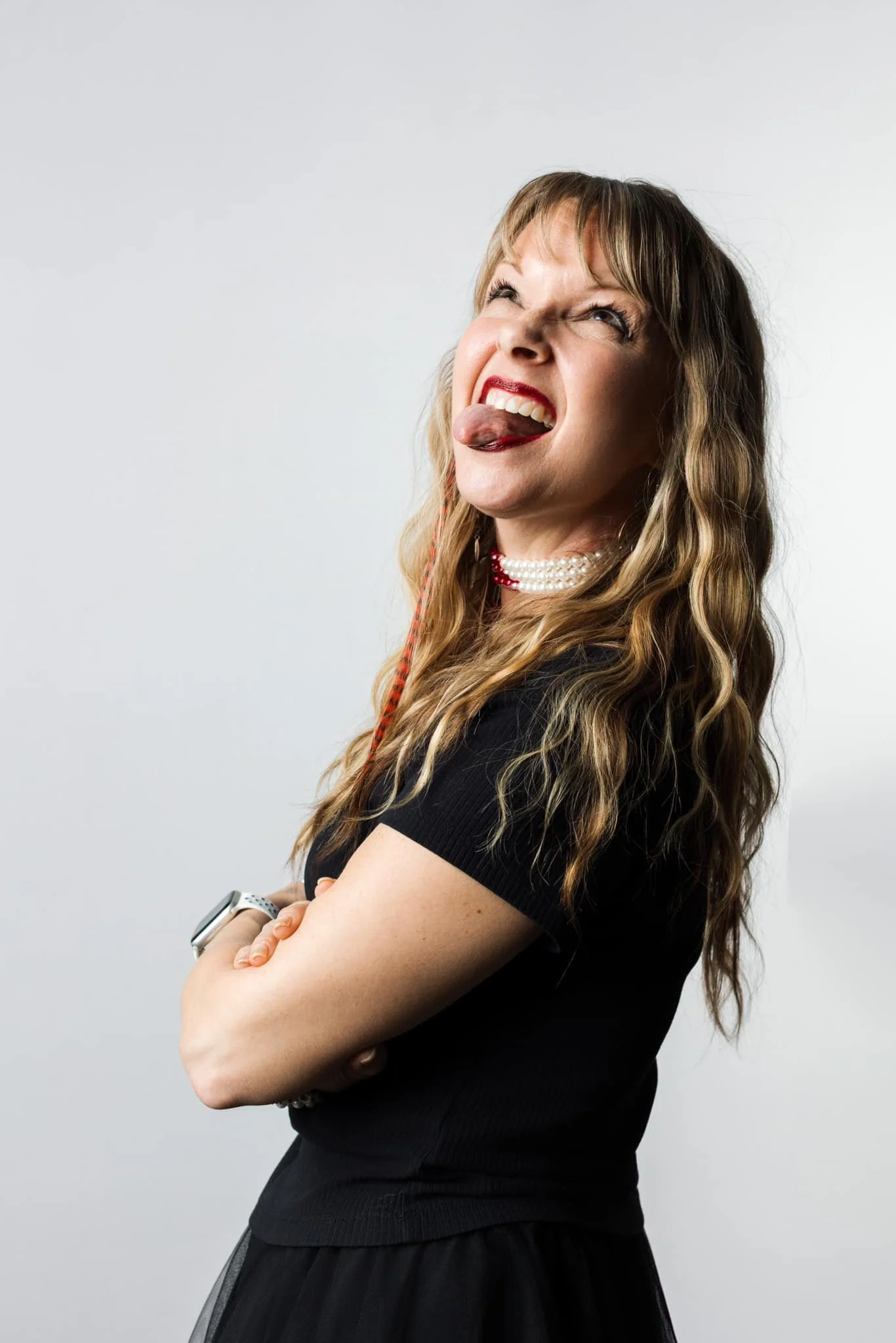 A woman with long wavy brown hair, wearing red lipstick, a pearl necklace, a black top, and a smartwatch, making a playful face with her tongue out and eyes squinted, standing against a plain white background.