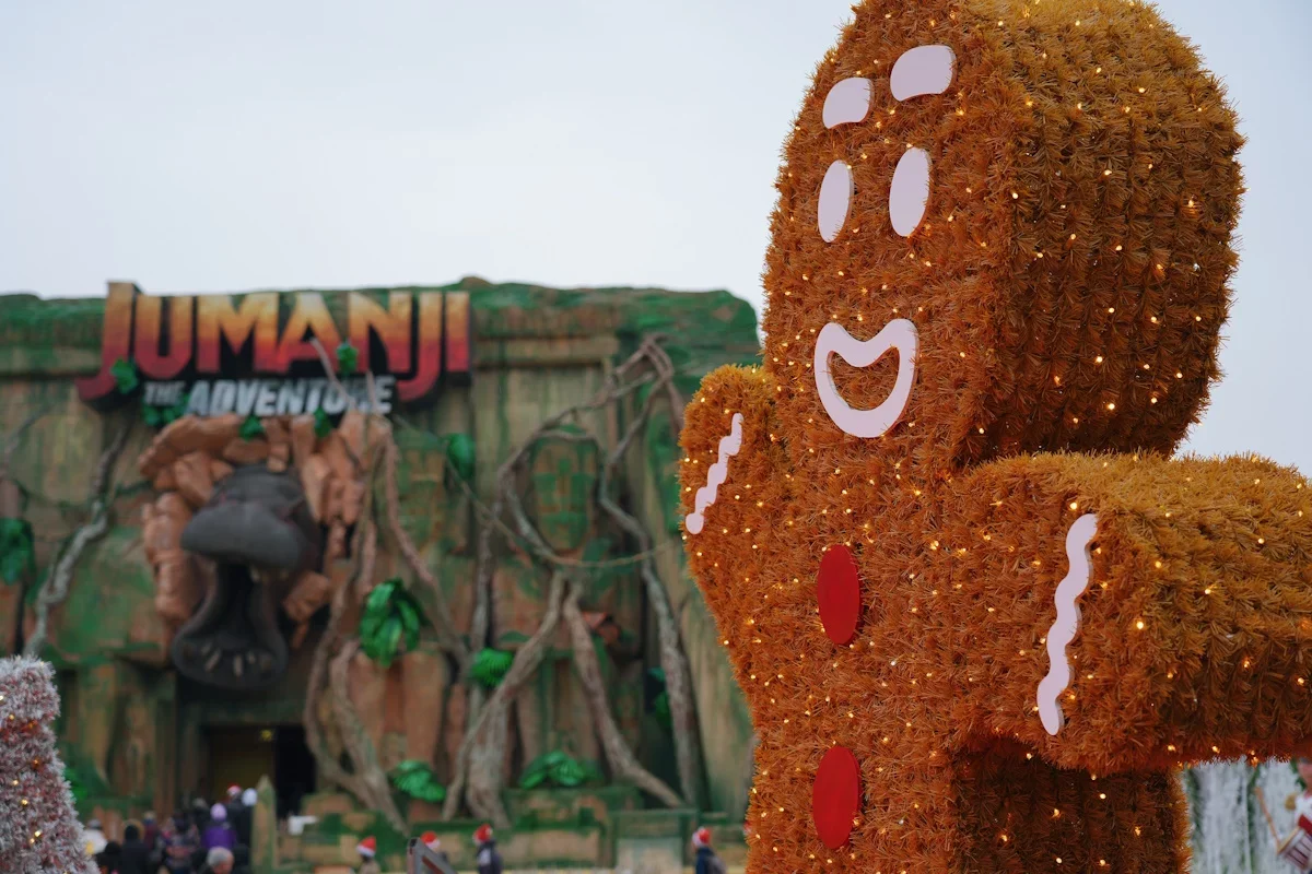 Decorative gingerbread man figure with smiling face, white eyes, and buttons, decorated with small lights, in front of the 'Jumanji: The Adventure' attraction entrance.