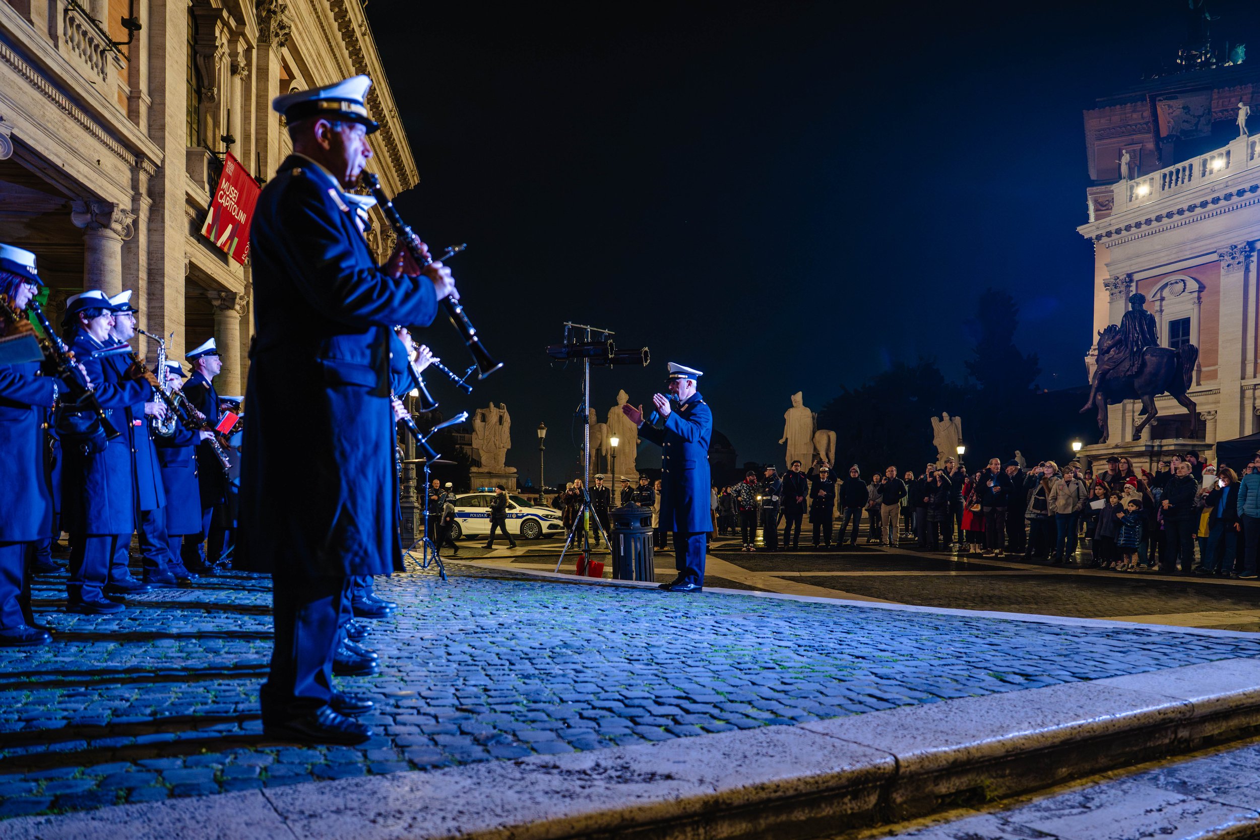 A nighttime outdoor concert featuring a military marching band playing instruments while standing on cobblestone pavement, with an audience watching. The backdrop includes historic architecture, statues, and a large building illuminated with lights.