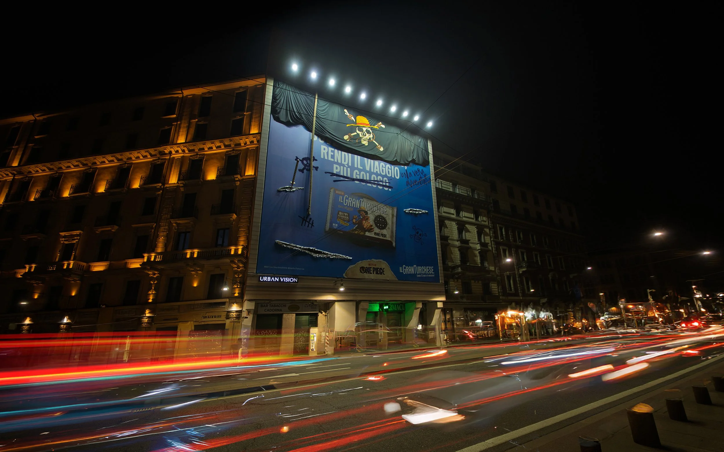Night view of buildings with illuminated windows and a large online advertisement banner featuring a cartoon pirate with a skull face and a hat, along with some text, on a city street with moving car lights.