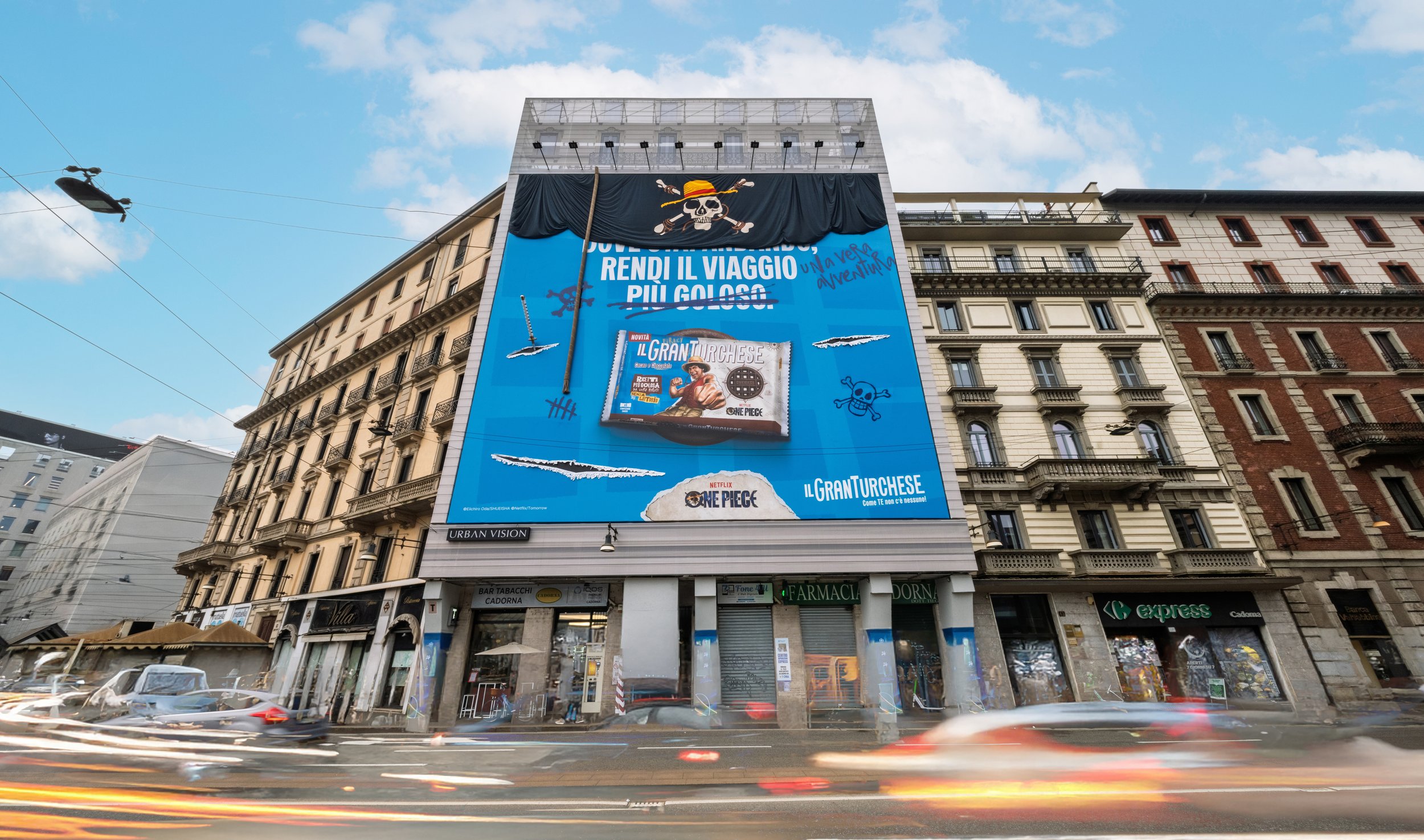 Night view of buildings with illuminated windows and a large online advertisement banner featuring a cartoon pirate with a skull face and a hat, along with some text, on a city street with moving car lights.
