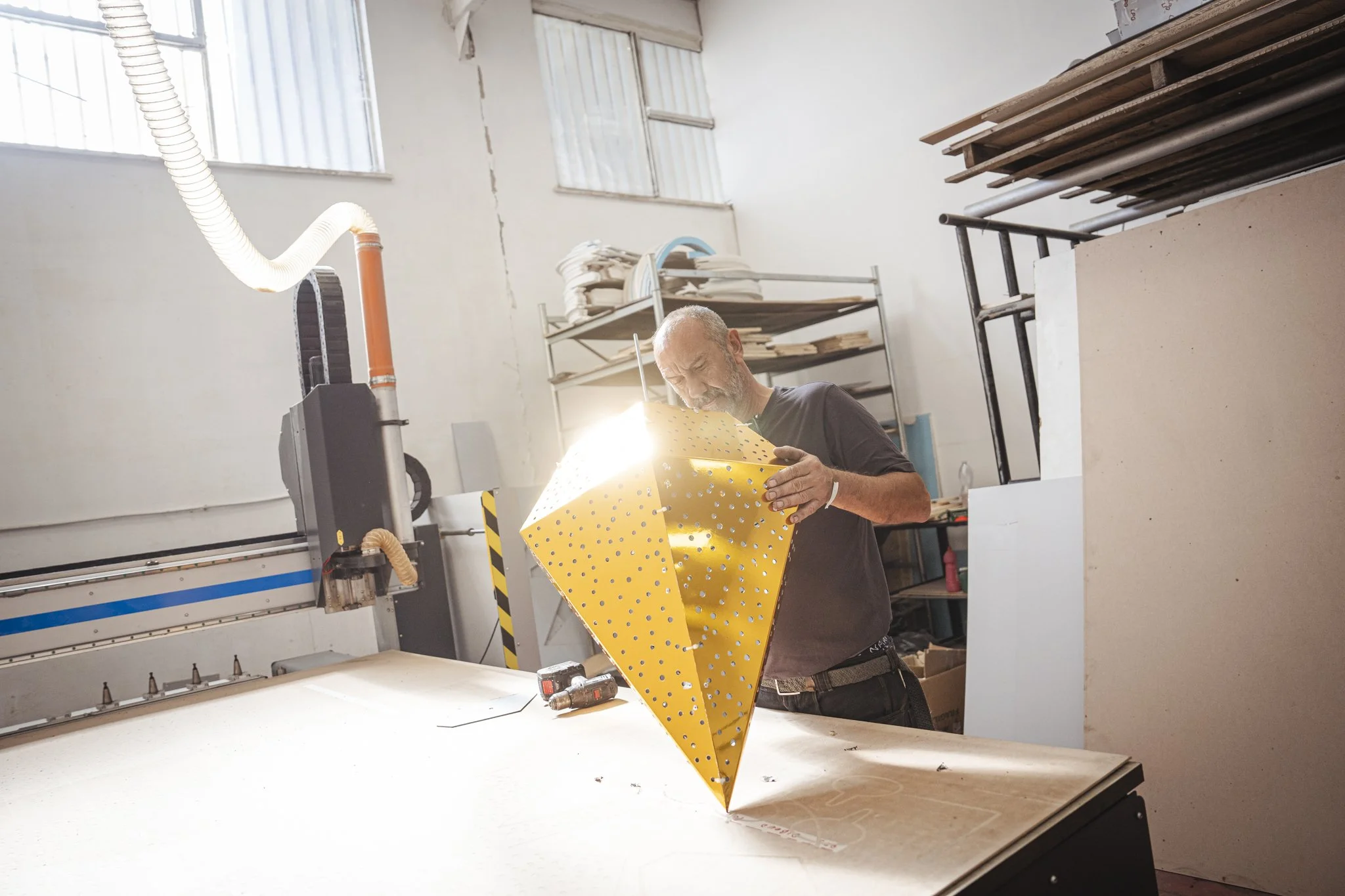 A man working in a workshop with a large machine, holding a yellow perforated sheet of metal, wearing a black shirt and glasses, with shelves and materials around him.
