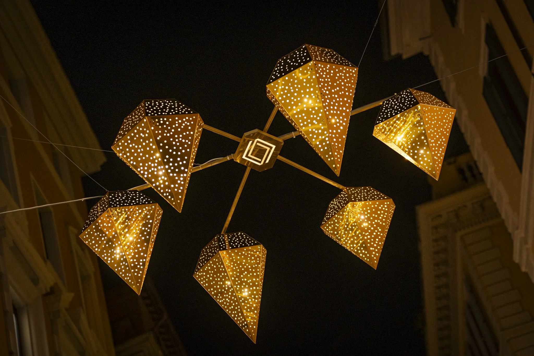 Decorative hanging lanterns with a perforated geometric pattern glowing in warm lights against a night sky, flanked by buildings.