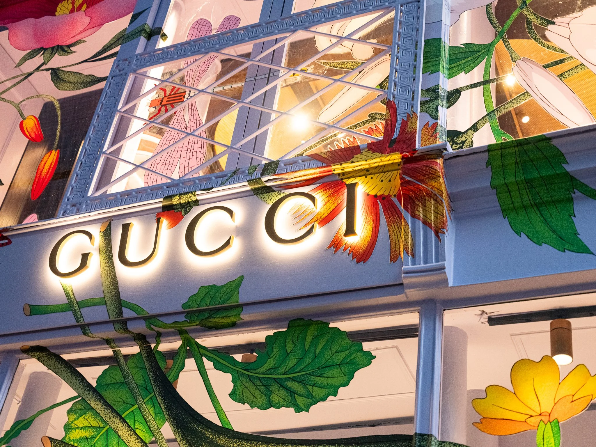 Close-up of a Gucci store sign with illuminated letters, decorated with floral and leaf illustrations, and a metal framework structure.