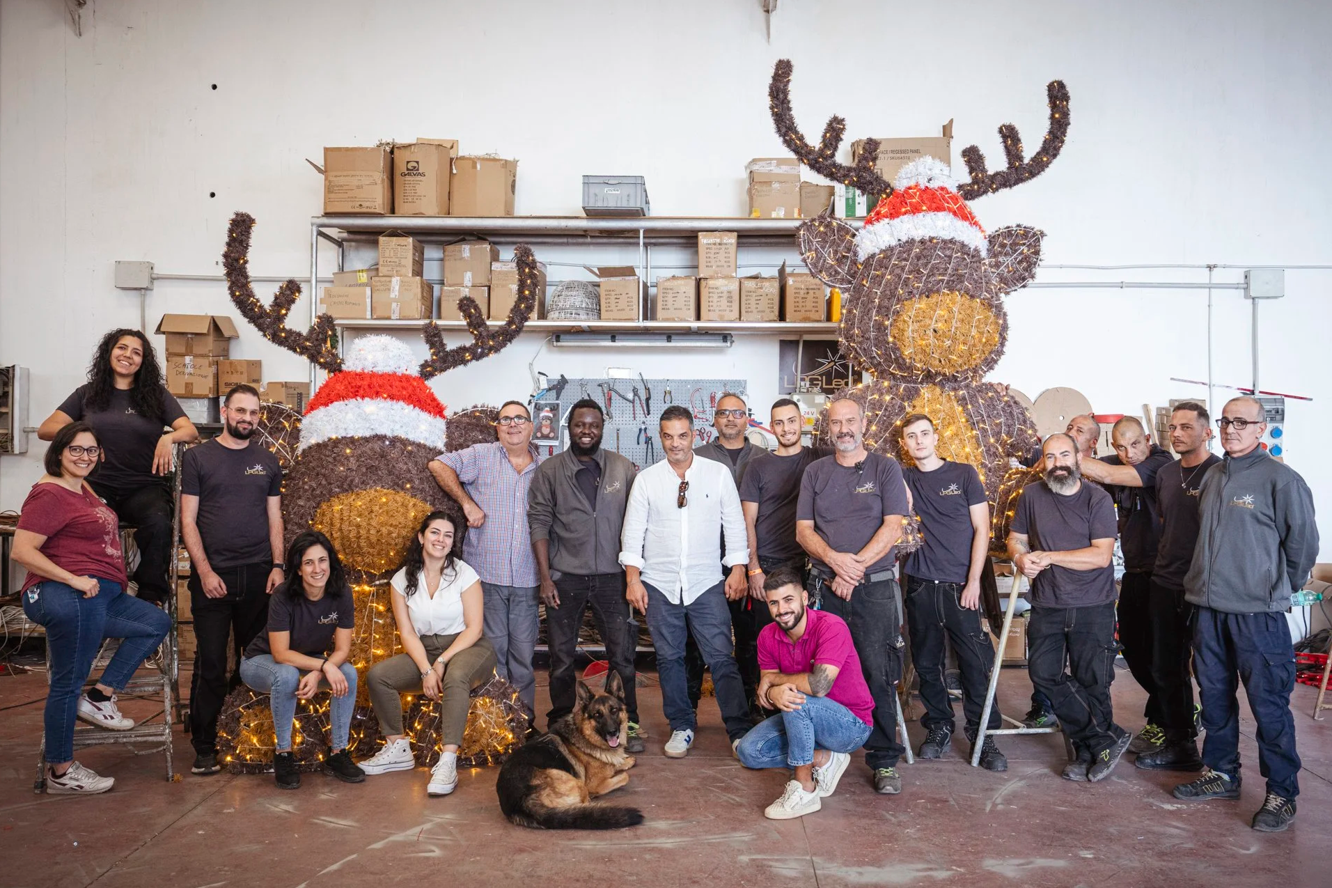 Group of people gathered in a workshop with two large reindeer decorations with Christmas hats and lights, and various tools and supplies in the background.