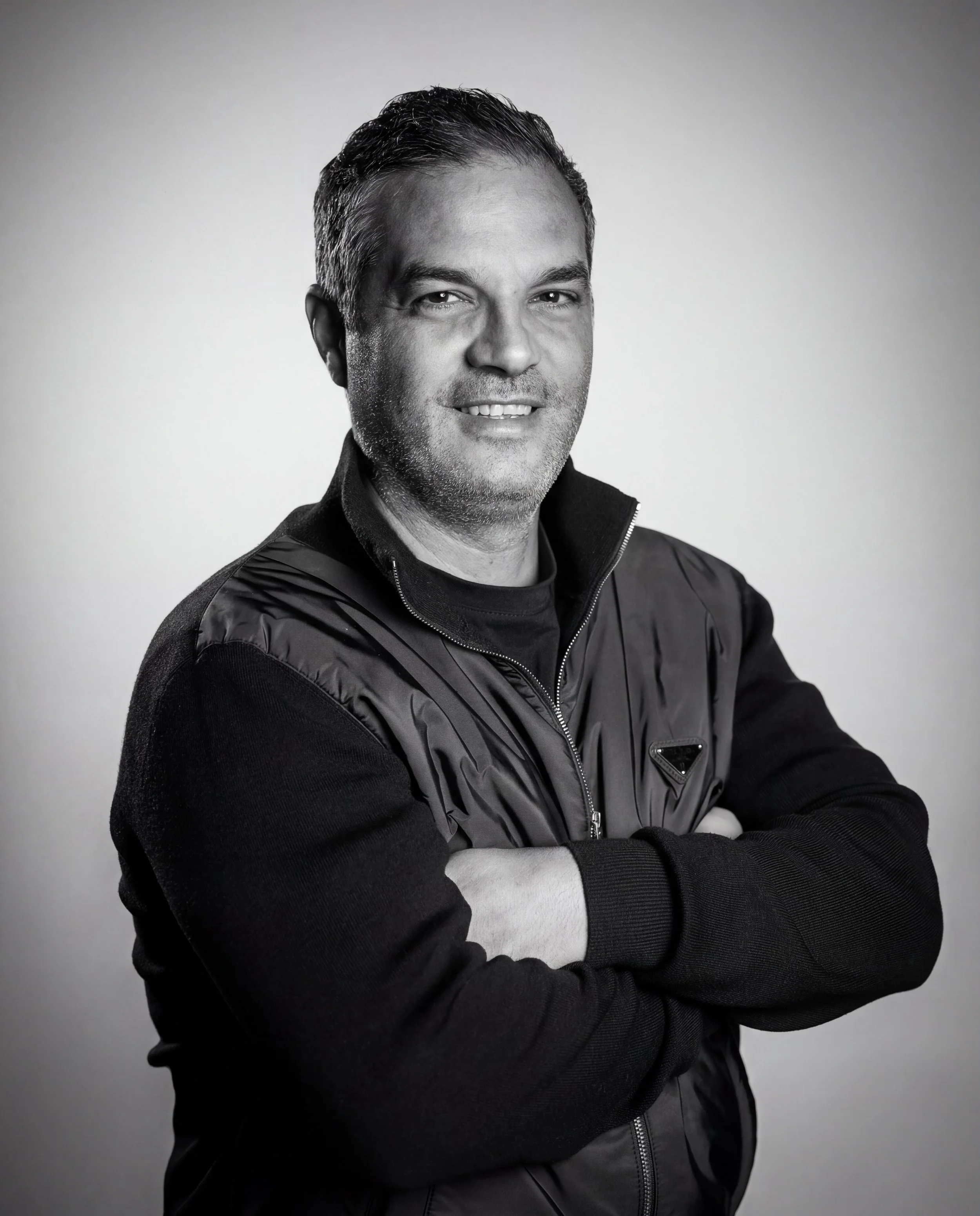 Black and white portrait of a middle-aged man with dark hair, smiling, wearing a dark jacket and a black shirt, with arms crossed in front of a plain light gray background.