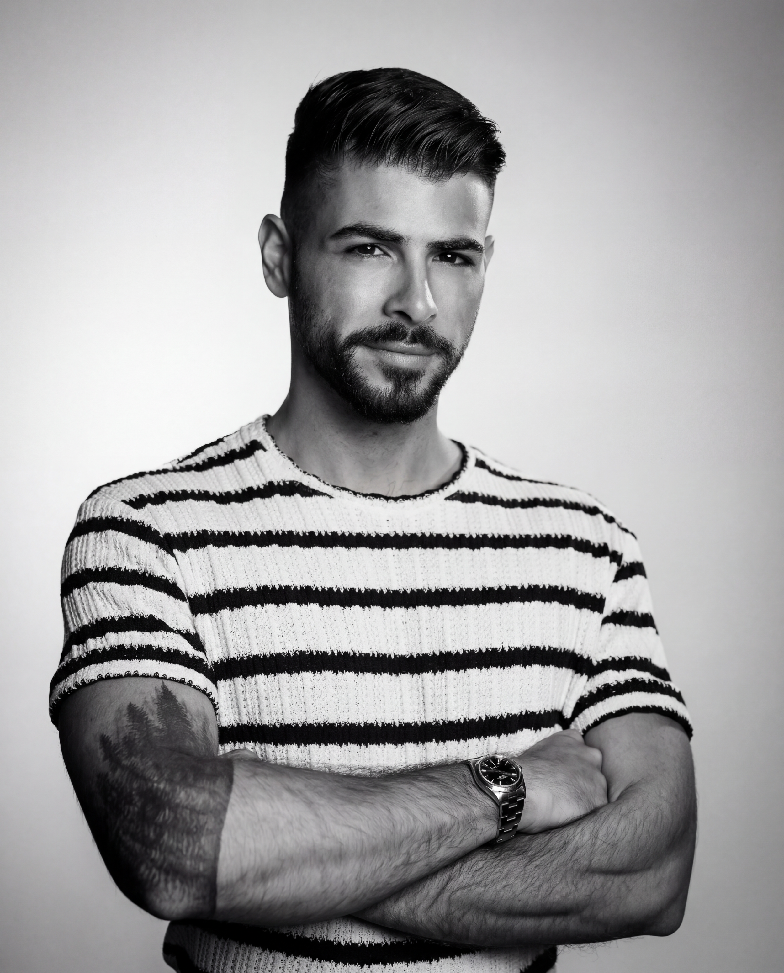 Black and white portrait of a young man with dark hair, a beard, and a tattoo on his left forearm, wearing a striped sweater and a wristwatch, with arms crossed, against a plain background.