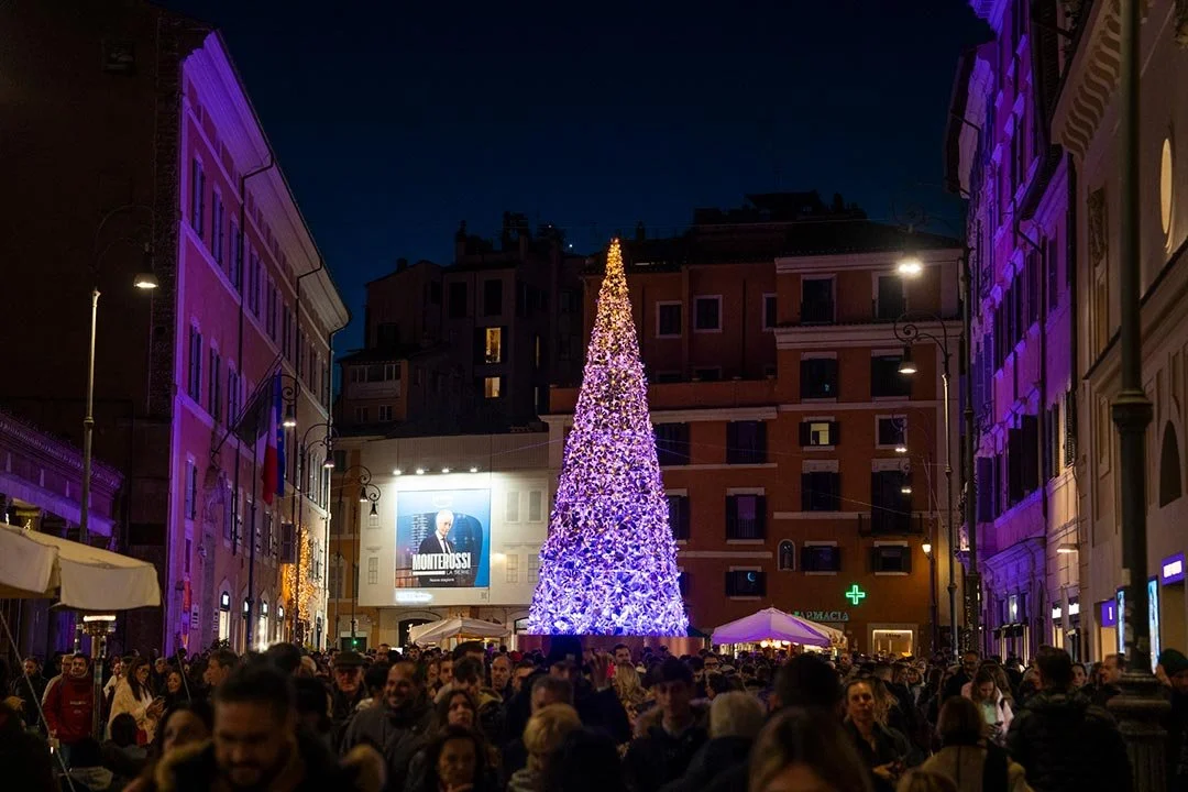 Crowd gathered around a large, decorated Christmas tree illuminated in purple and yellow lights in a city square at night.