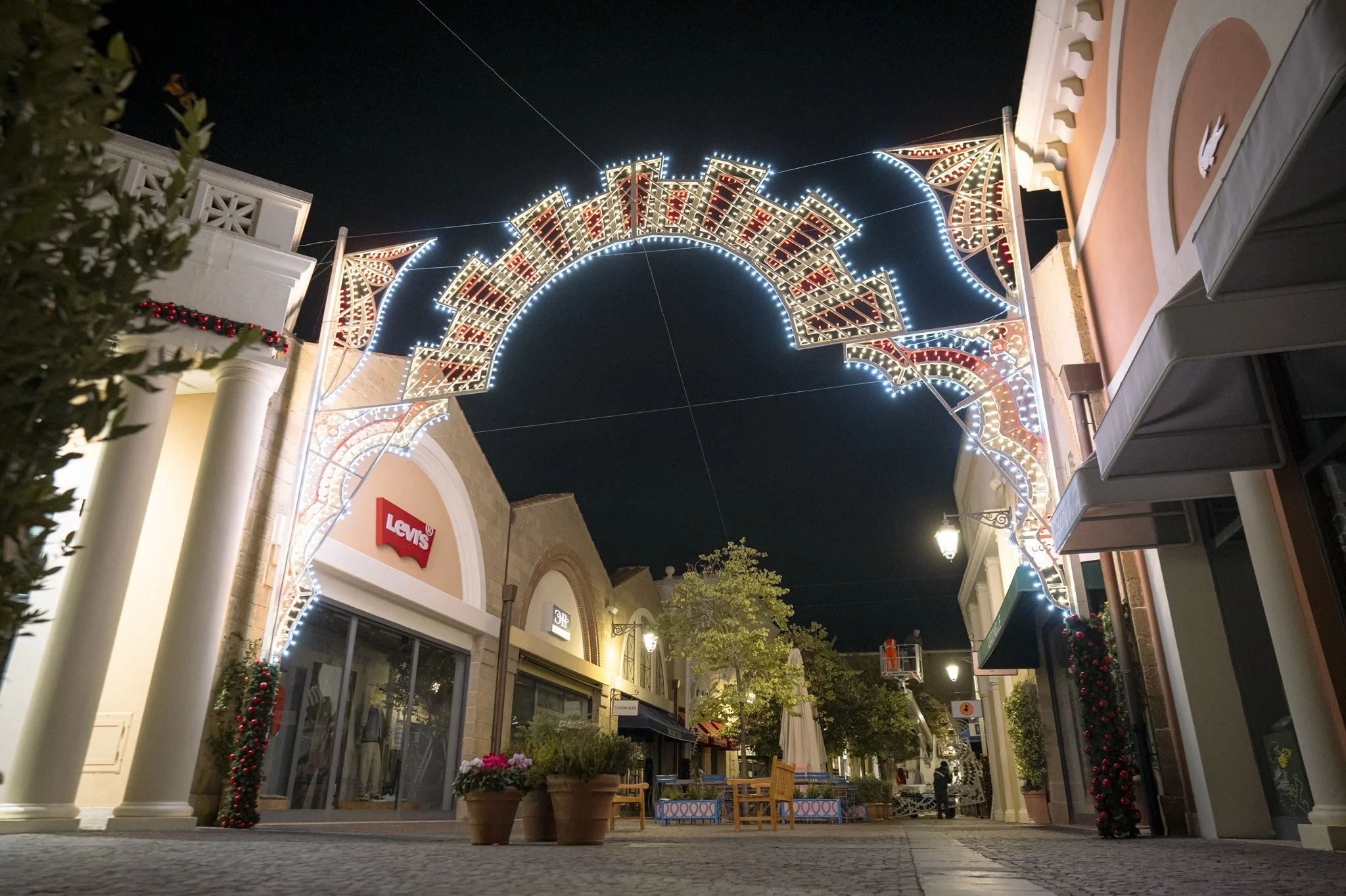 Night view of a shopping area with decorative lights forming a festive arch, storefronts including Levi's, and street lamps illuminating the scene.'}