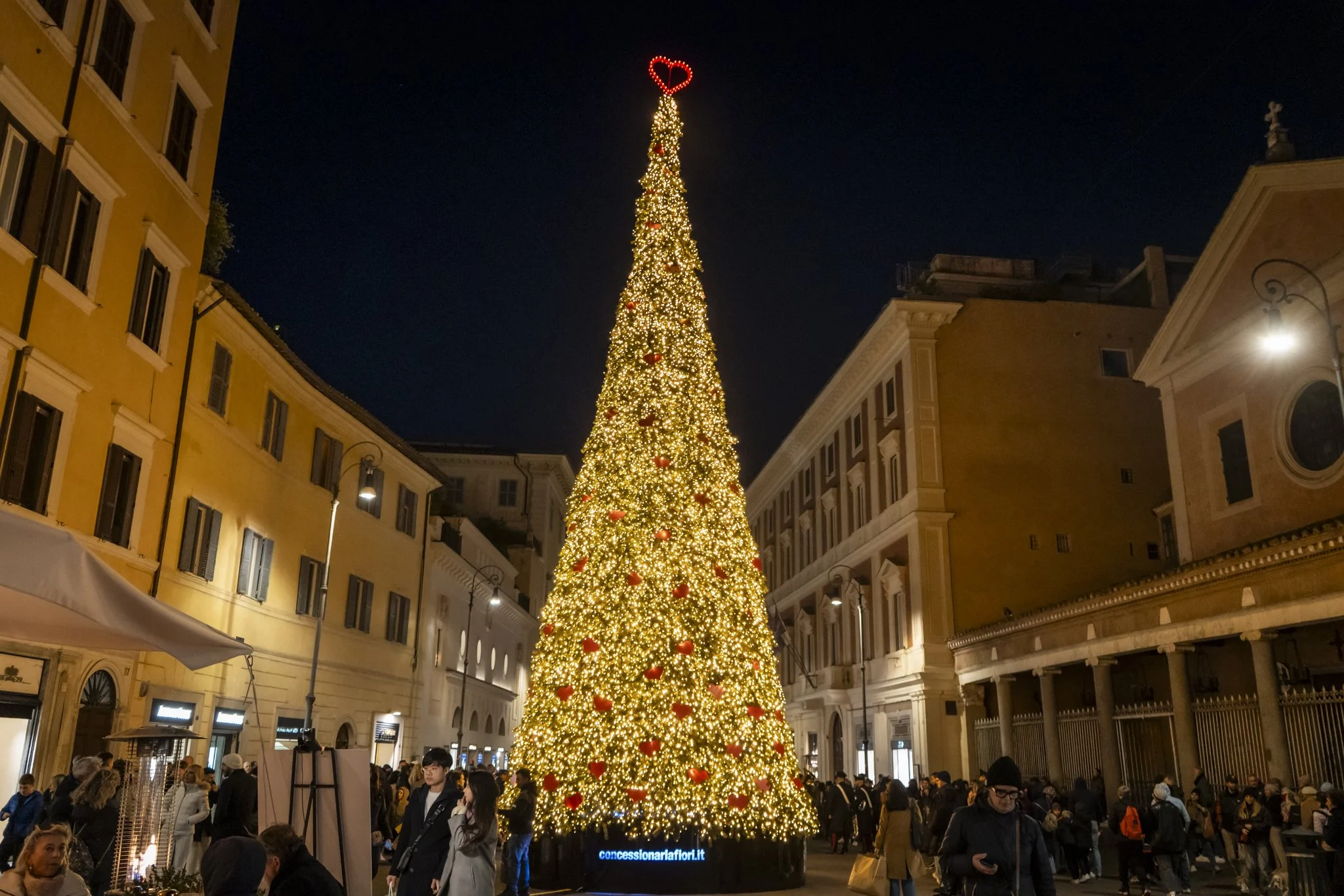 Large Christmas tree decorated with lights and red ornaments, topped with a red heart-shaped decoration, surrounded by a crowd of people during a night celebration in an urban setting.