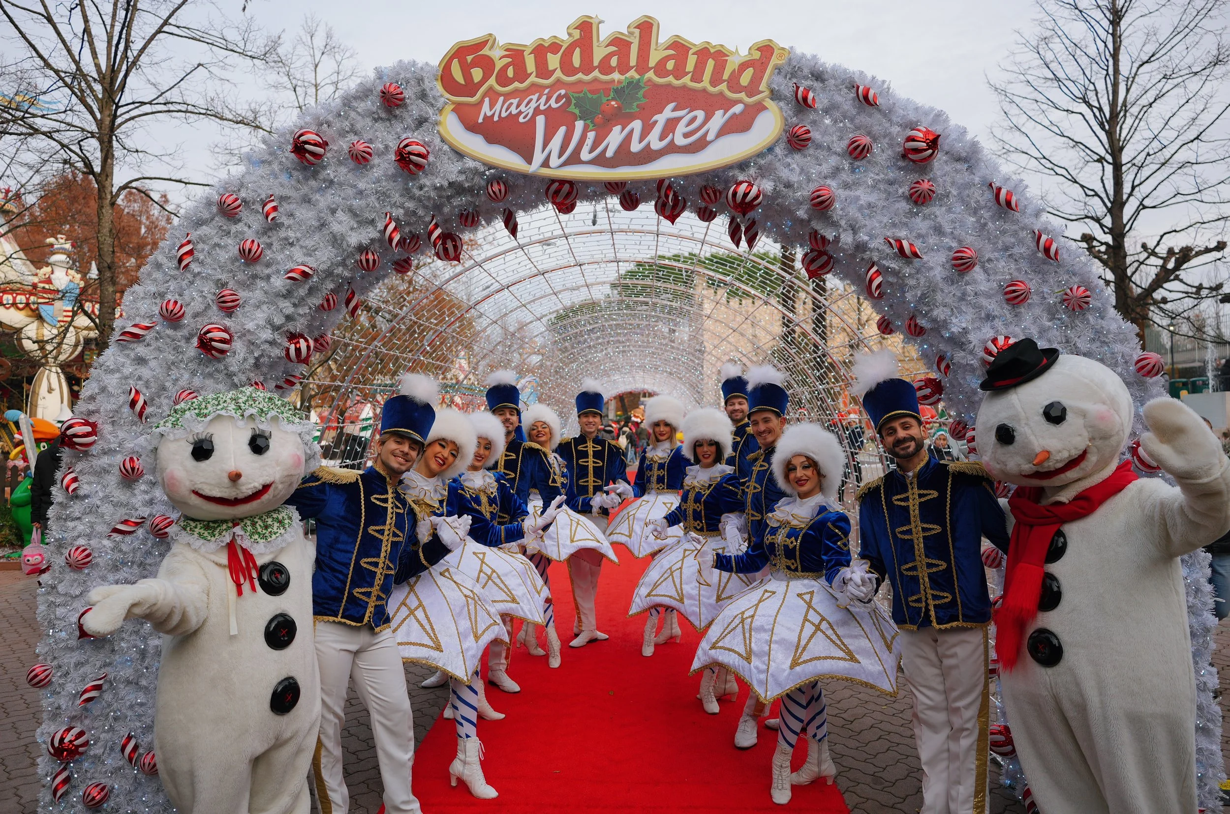 A festive parade scene with performers in colorful costumes and mascot characters dressed as snowmen, under an archway decorated with white and red ornaments. The arch has a sign that reads 'Gardaland Magic Winter' and the sky is overcast with trees in the background.