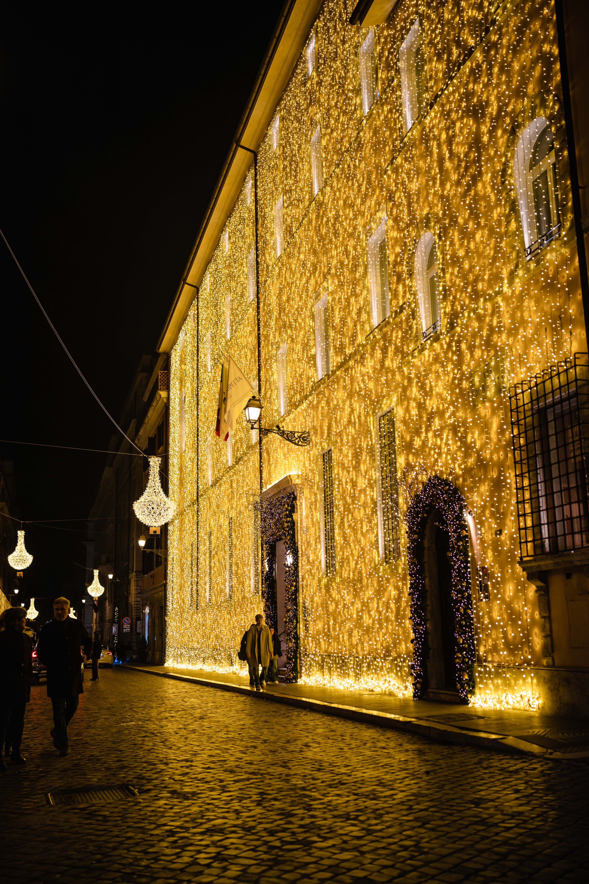 A street illuminated with festive golden Christmas lights hanging from a building and decorative chandeliers suspended above the cobblestone street at night.