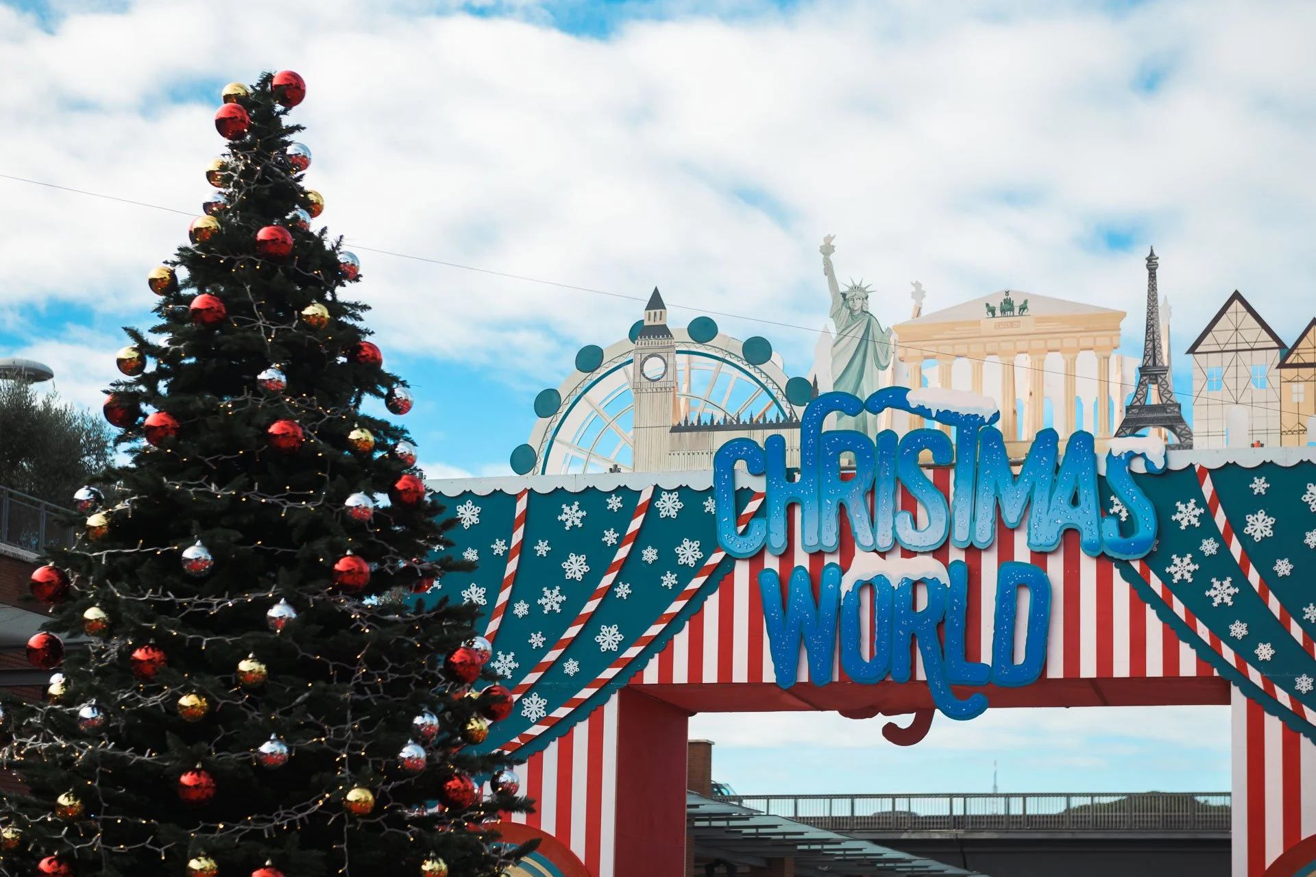 A decorated Christmas tree with red and gold ornaments and lights, set against a backdrop of amusement park signs featuring iconic landmarks such as the Statue of Liberty, Big Ben, the Eiffel Tower, and the Parthenon, with a festive sign that reads 'Christmas Around the World.'