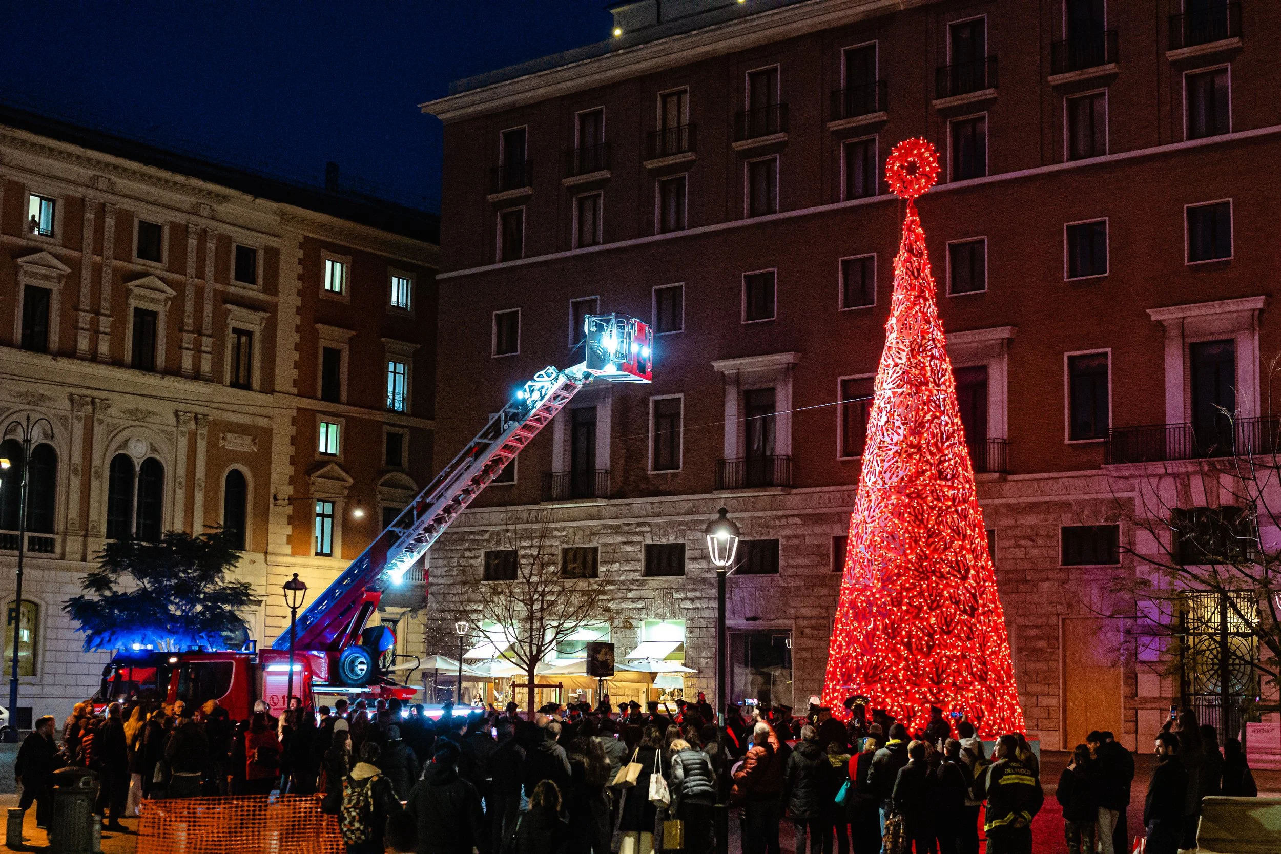 A large crowd gathered in an urban plaza at night, watching a fire truck with an extended ladder illuminated by bright lights. The ladder is reaching towards a tall, illuminated holiday tree decorated with red lights, in front of multi-story buildings with lit windows.