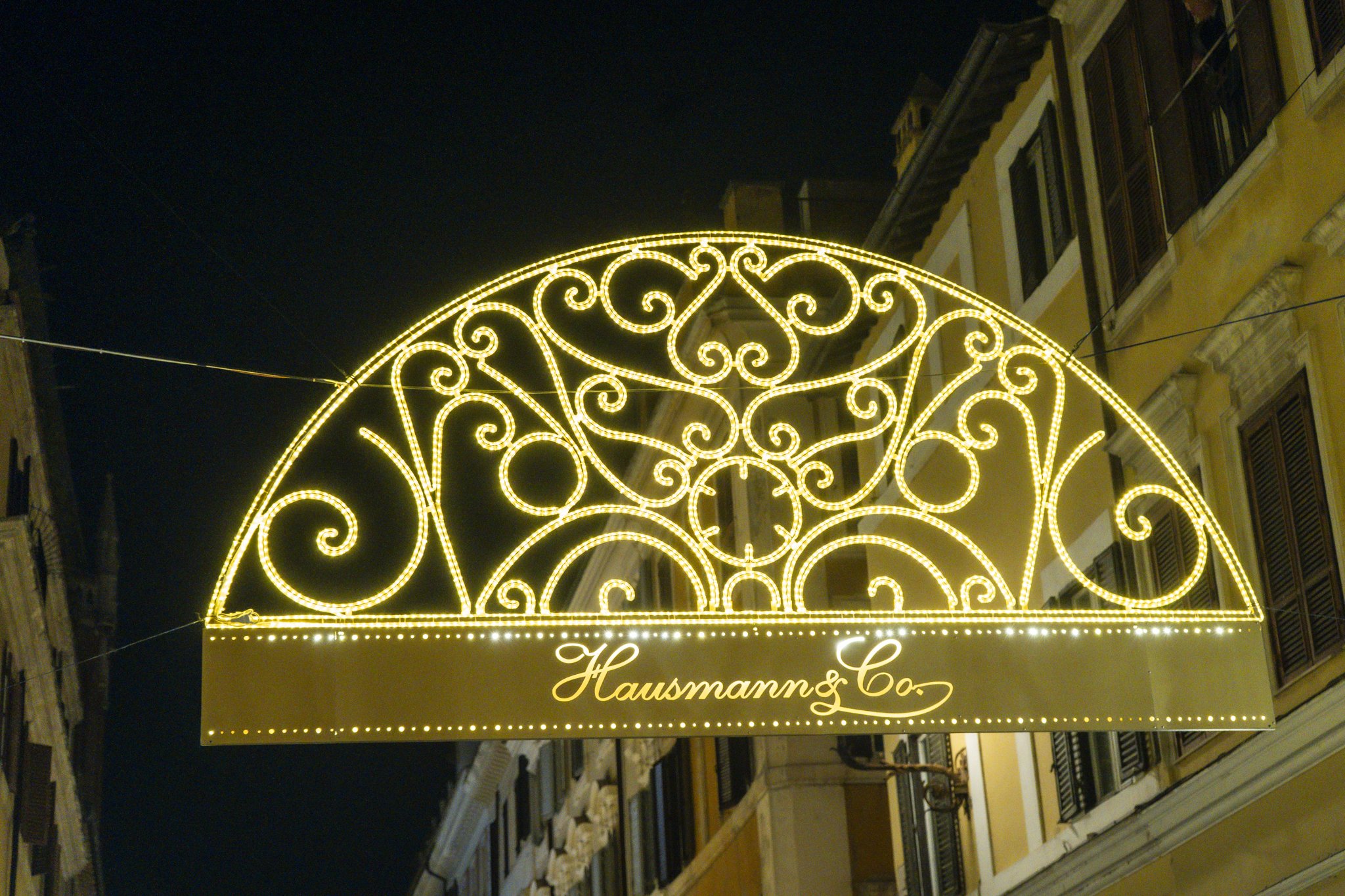 Illuminated decorative sign with intricate gold swirling patterns hanging over a street at night, with the words 'Hausmann & Co.' written at the bottom.