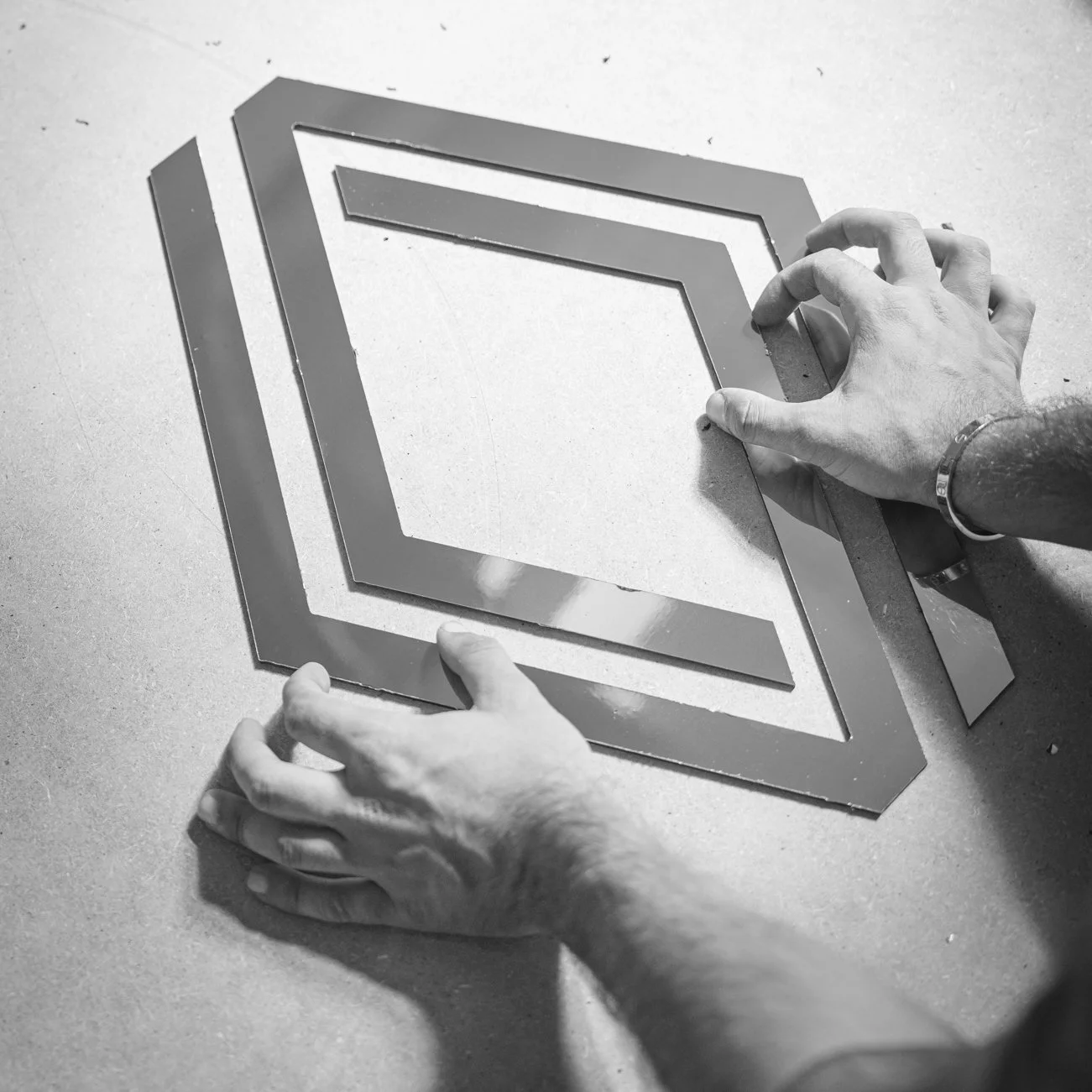 A person assembling a metal frame, possibly a stencil, on a flat surface in black and white.