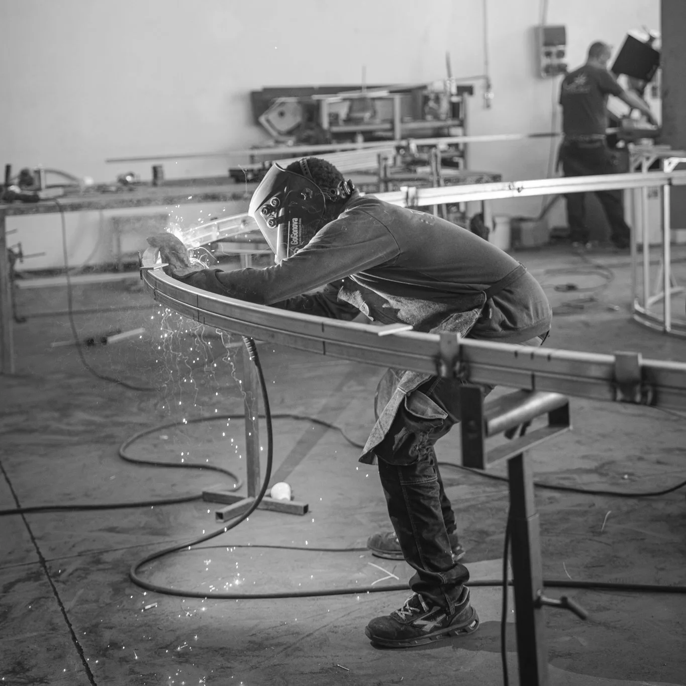Person welding metal in a workshop, wearing a welding helmet with sparks flying, while another person works at a machine in the background.