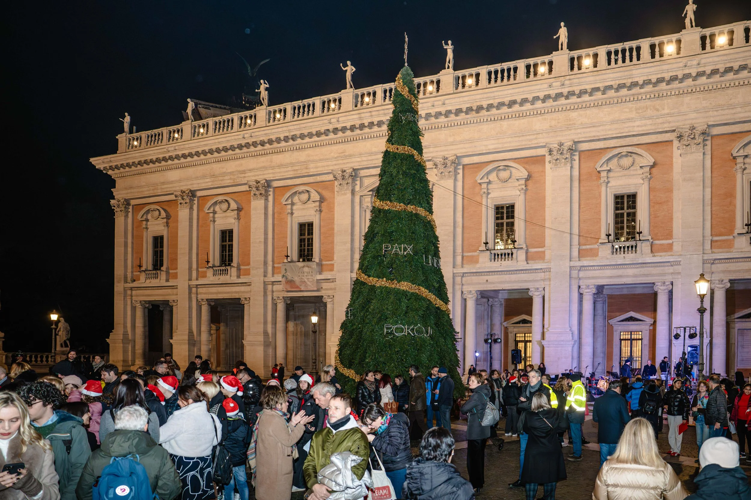 Nighttime scene in a large plaza with a crowd of people, many wearing Santa hats, gathered around a decorated Christmas tree in front of a historic building with statues on its roof and illuminated windows.