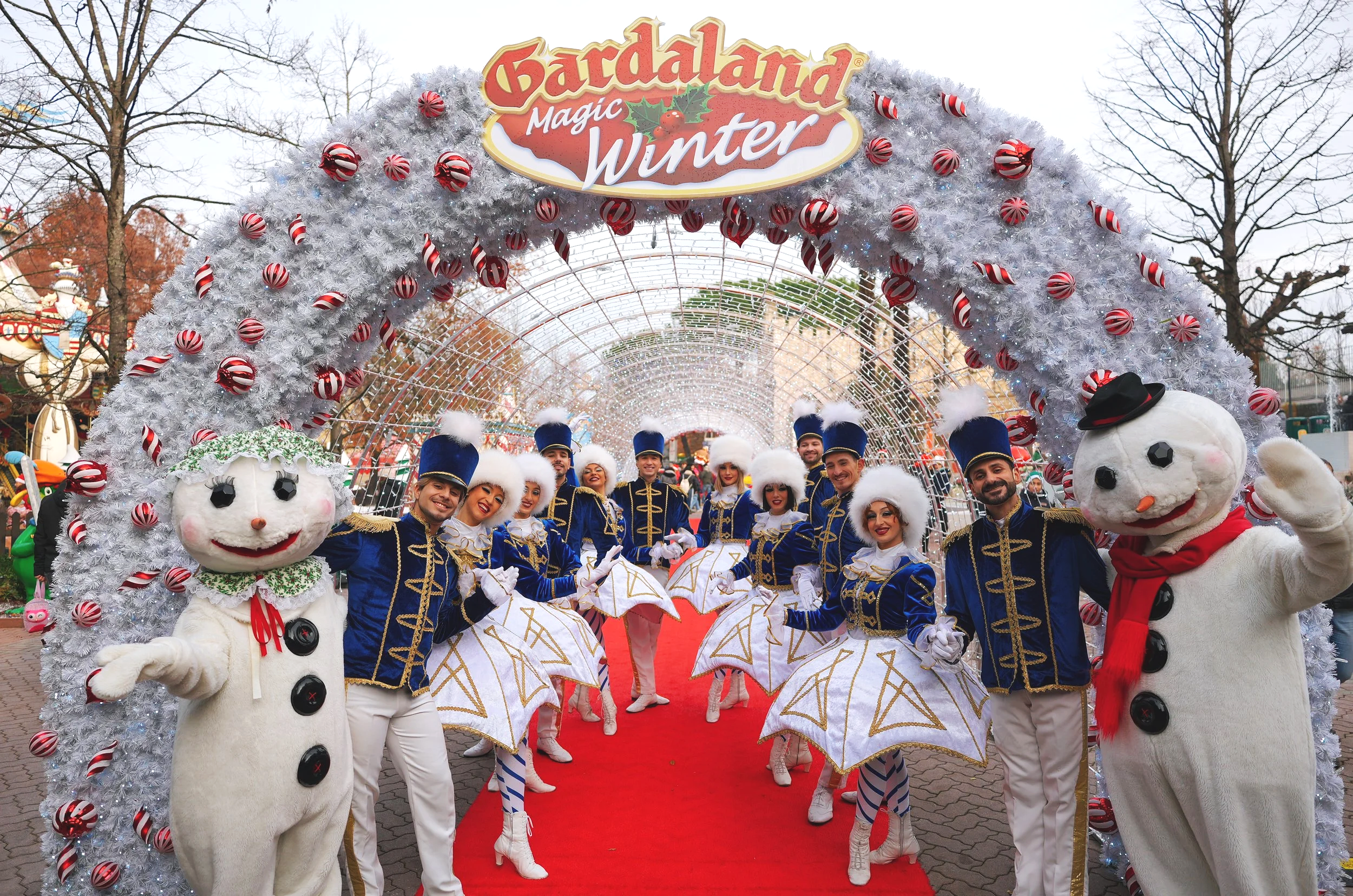 A festive parade scene with performers in colorful costumes and mascot characters dressed as snowmen, under an archway decorated with white and red ornaments. The arch has a sign that reads 'Gardaland Magic Winter' and the sky is overcast with trees in the background.