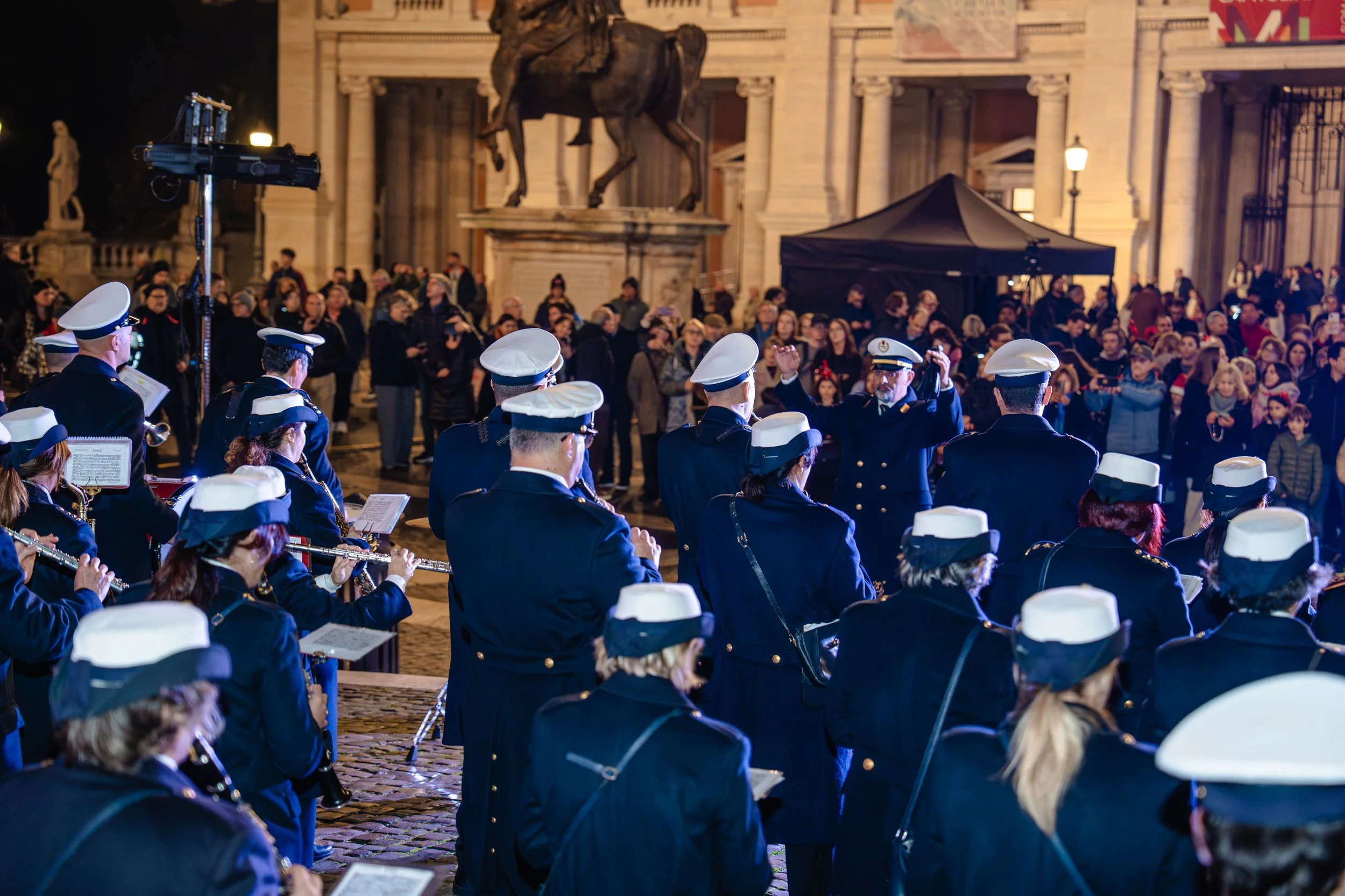 A nighttime scene of a crowd of people watching a band of musicians dressed in military-style uniforms with white hats, performing outdoors in front of a historic building with large columns and statues. The setting appears festive, possibly a public