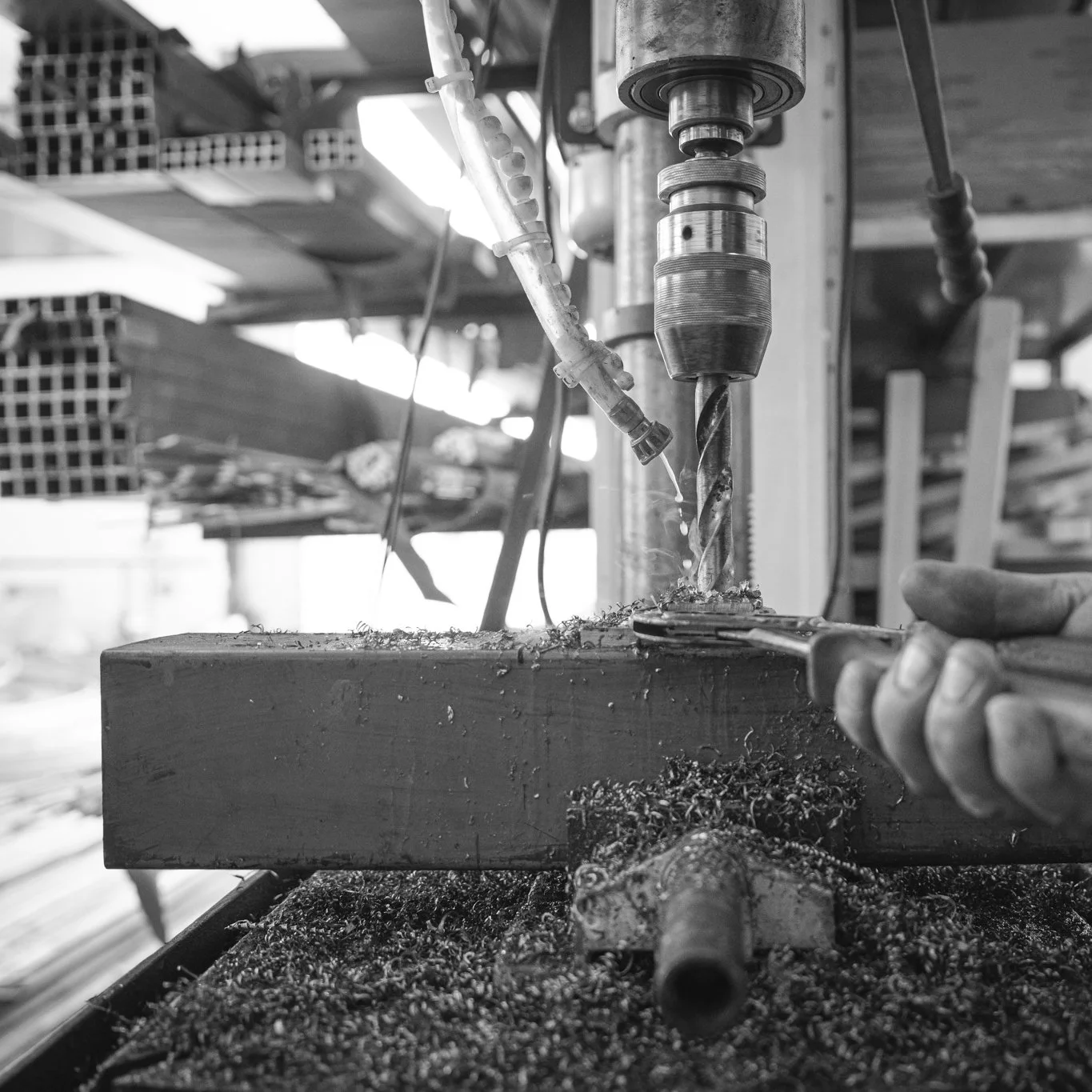 A person operating a drill press in a woodworking shop, drilling a hole into a piece of wood with wood shavings and debris around.