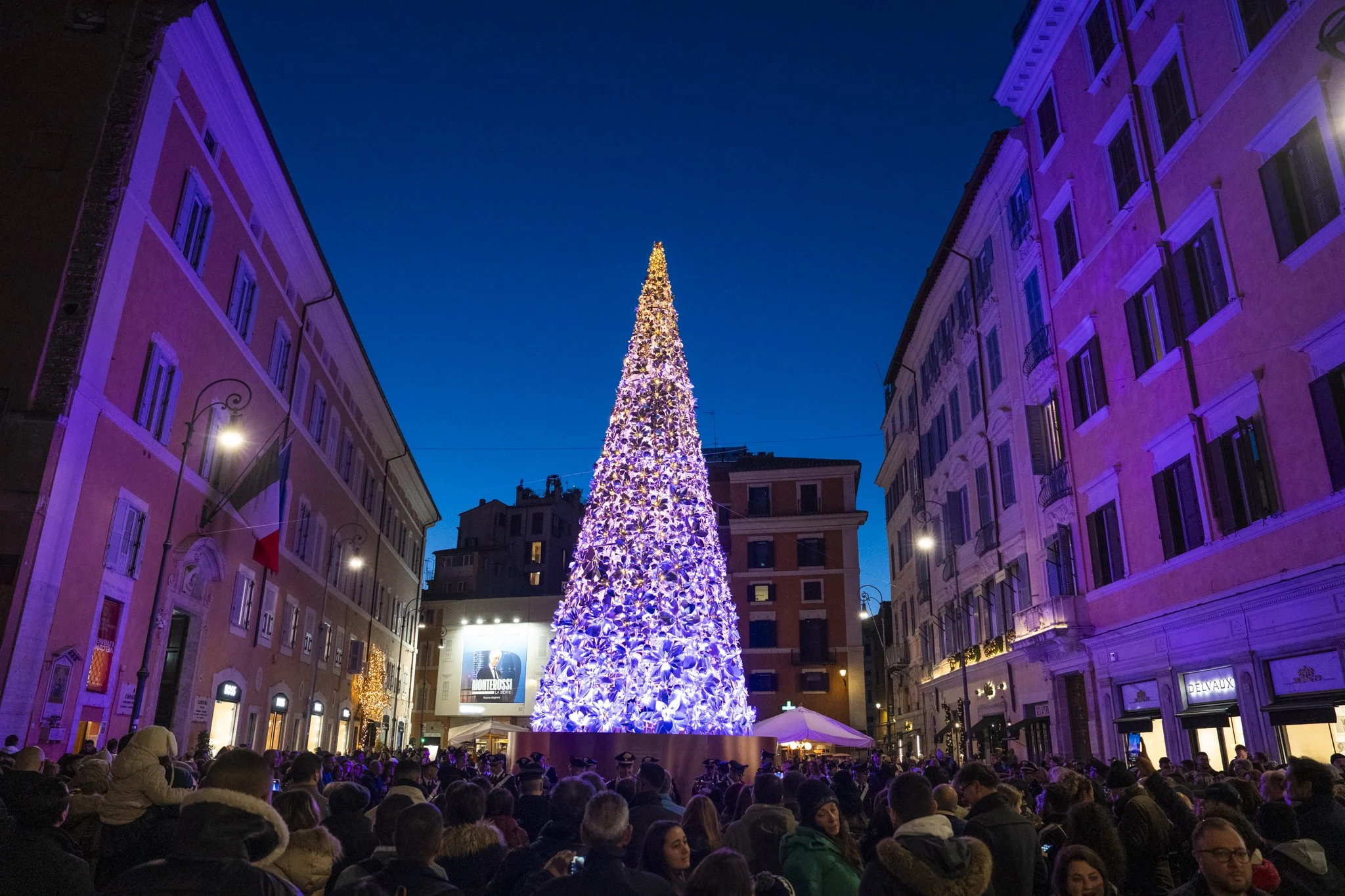 Large Christmas tree decorated with lights and red ornaments, topped with a red heart-shaped decoration, surrounded by a crowd of people during a night celebration in an urban setting.