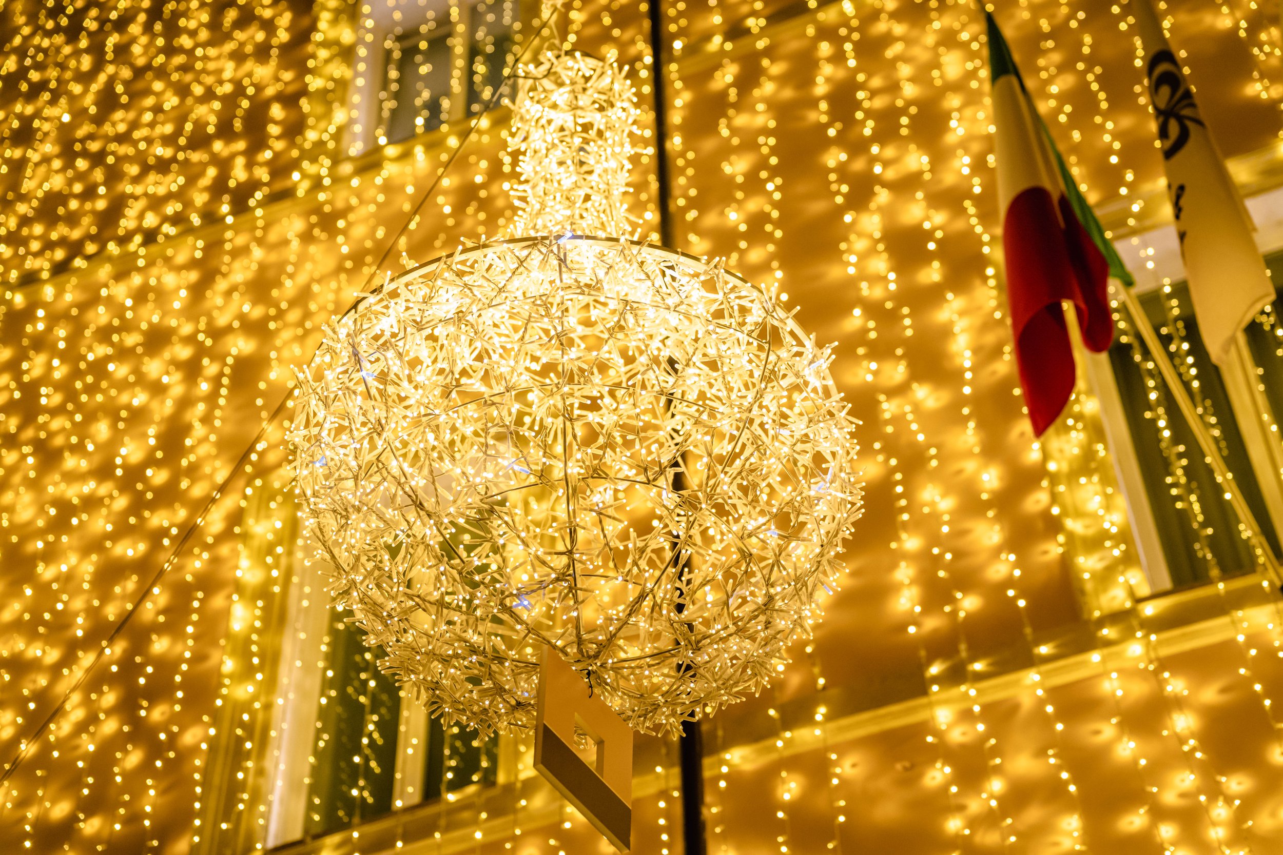 A street illuminated with festive golden Christmas lights hanging from a building and decorative chandeliers suspended above the cobblestone street at night.
