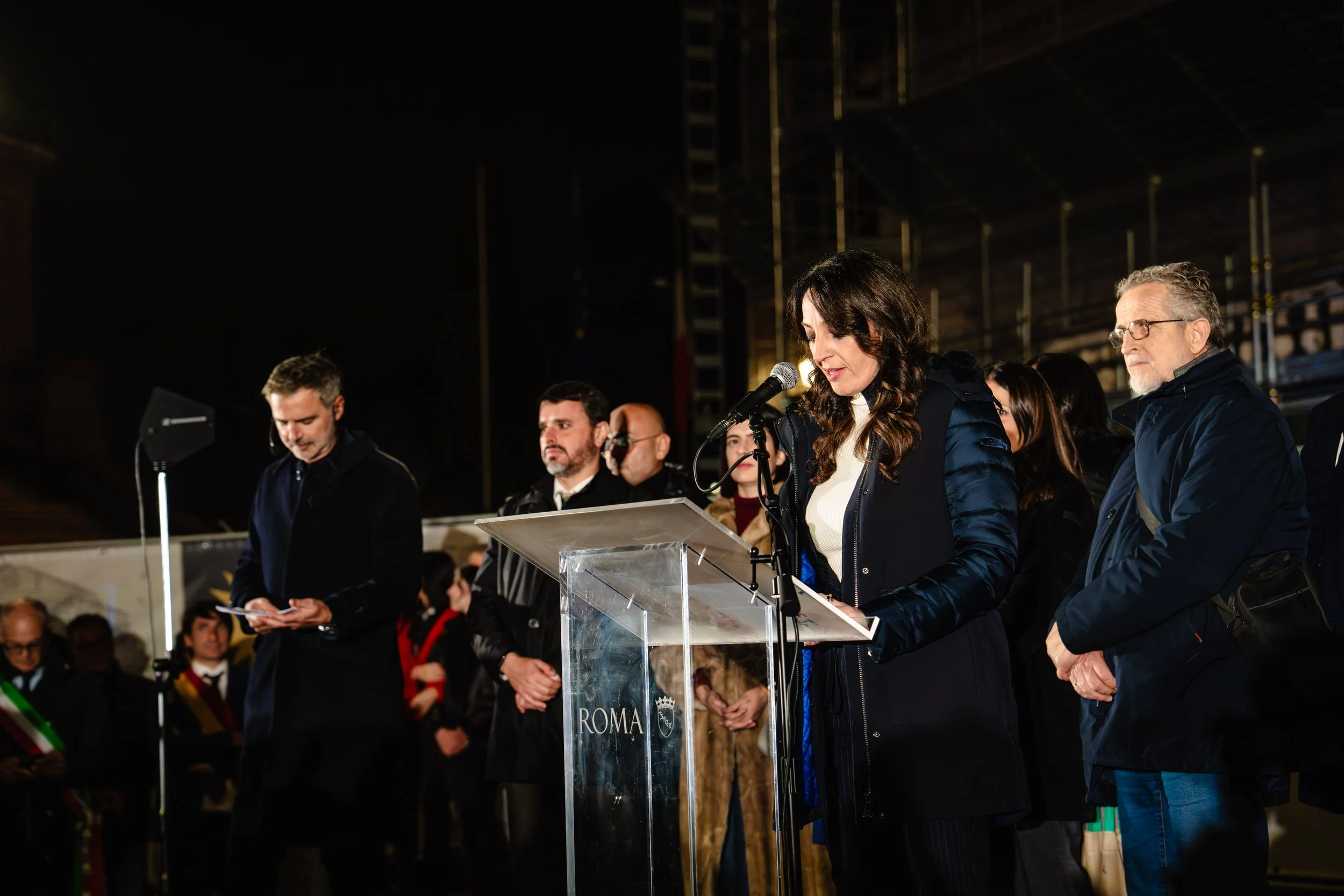 A group of people gathered at a podium with a woman speaking into a microphone, in an indoor event or rally setting.