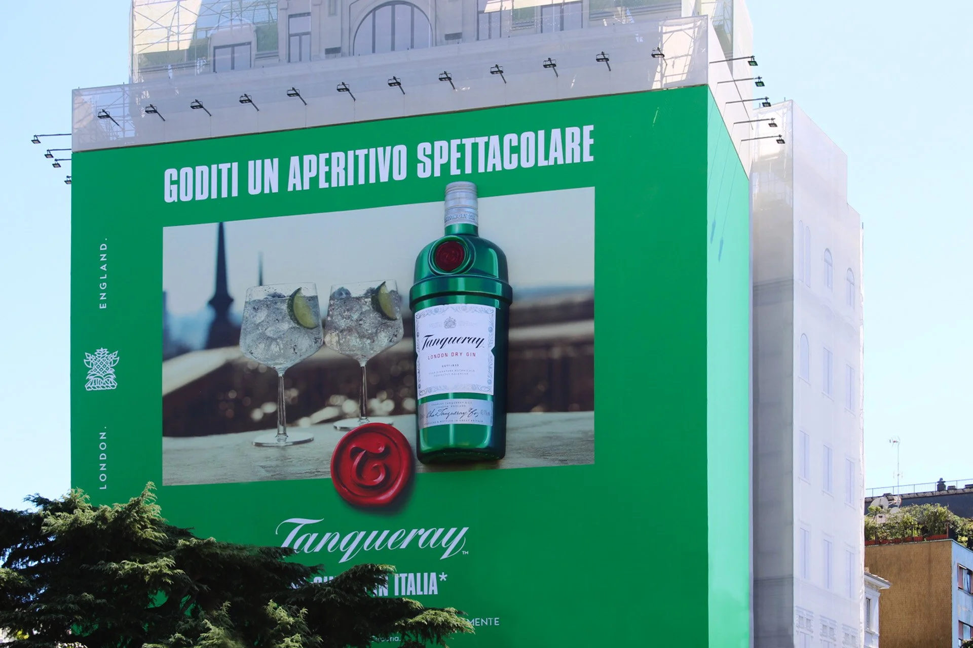 Billboard advertising Tanqueray London Dry Gin featuring a green bottle, two glasses with ice and lime wedges, and the message in Italian about enjoying a spectacular aperitif.